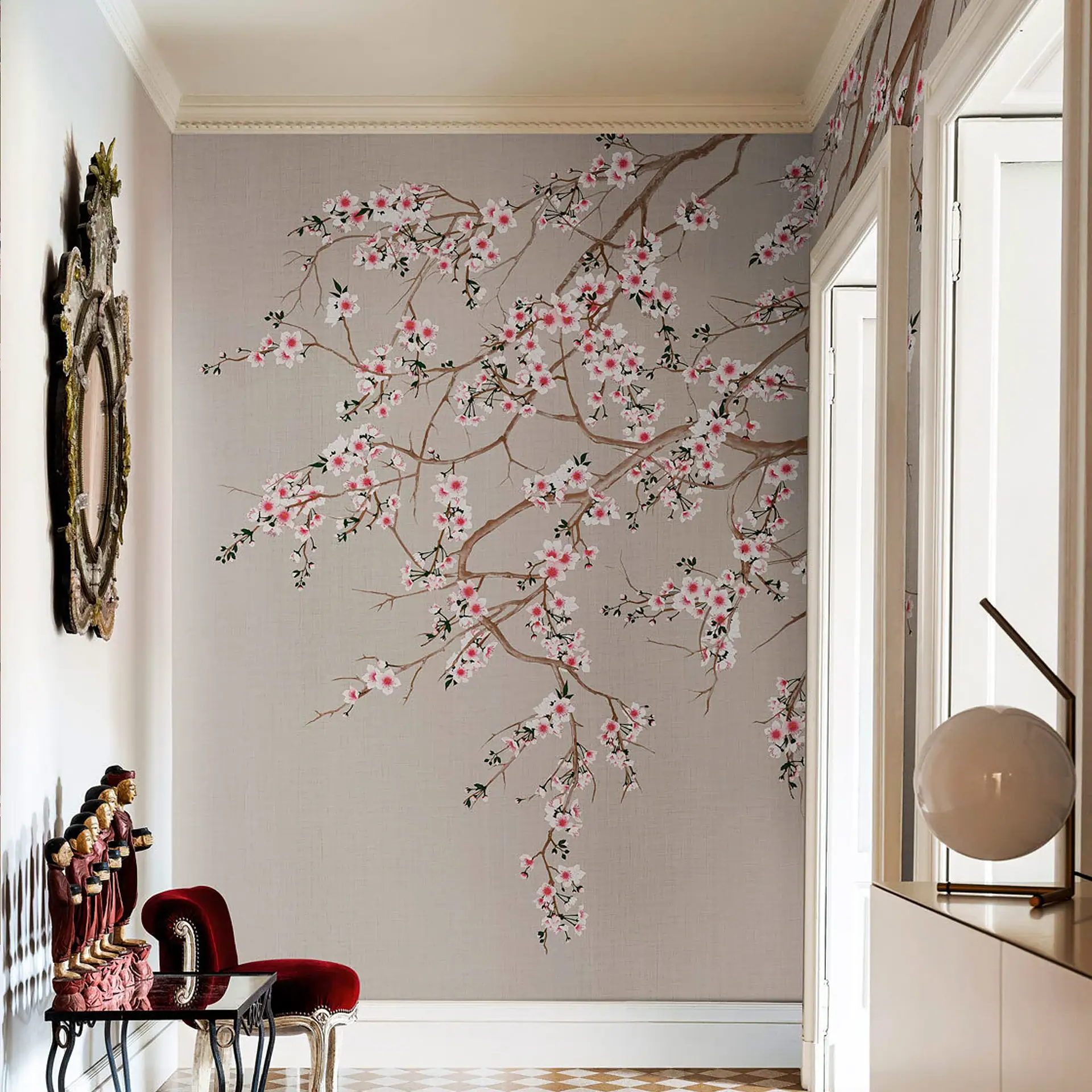 A luxurious hallway with light grey cherry blossom wallpaper, an ornate mirror, a red velvet chair, wooden figurines, and a modern spherical lamp on a white cabinet.