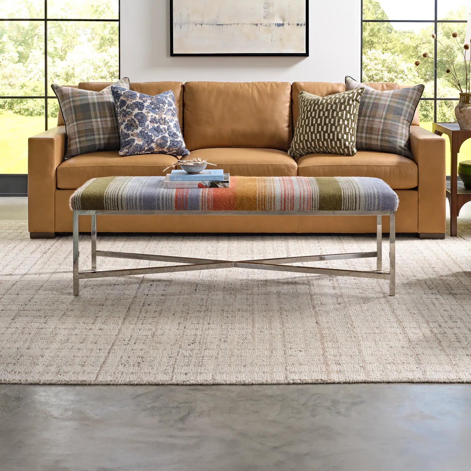 A living room featuring a brown leather sofa, a colorful striped bench, and a light textured rug. Large windows reveal green trees, and abstract art hangs above the sofa.
