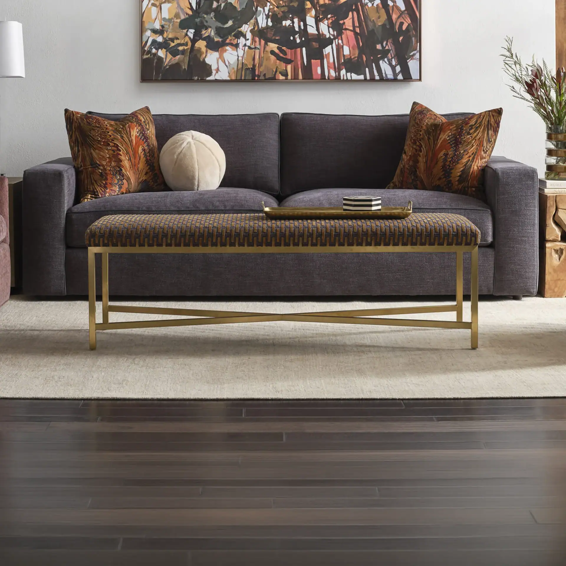 A living room with a dark gray sofa, patterned pillows, a geometric upholstered bench with a brass frame, and abstract art on the wall. Dark hardwood floors are in the foreground.