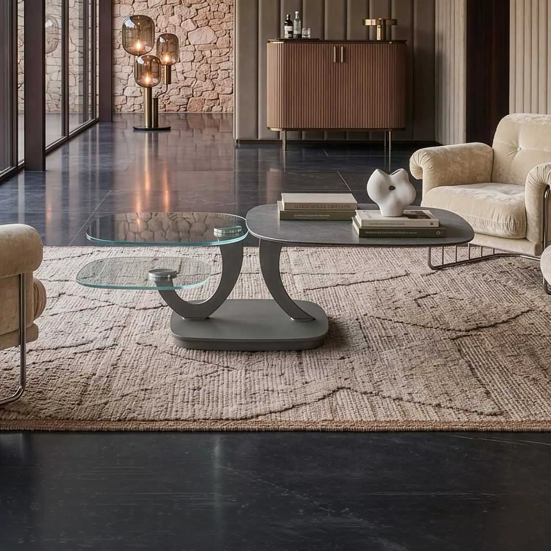 A modern living room with a gray and glass coffee table on a textured rug, flanked by cream armchairs. A fluted cabinet and a three-light floor lamp are in the background.