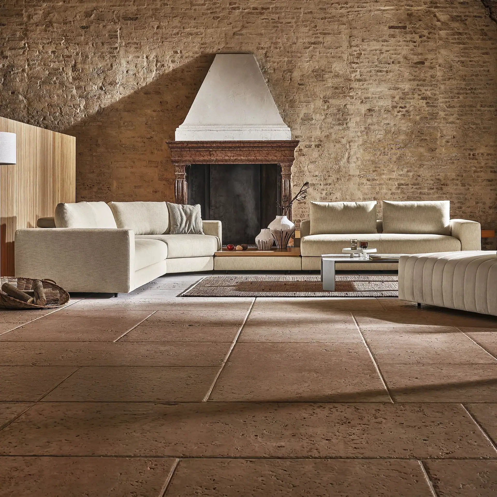 A living room with exposed brick walls, a large fireplace, a light beige sectional sofa, a coffee table, and stone tile flooring.