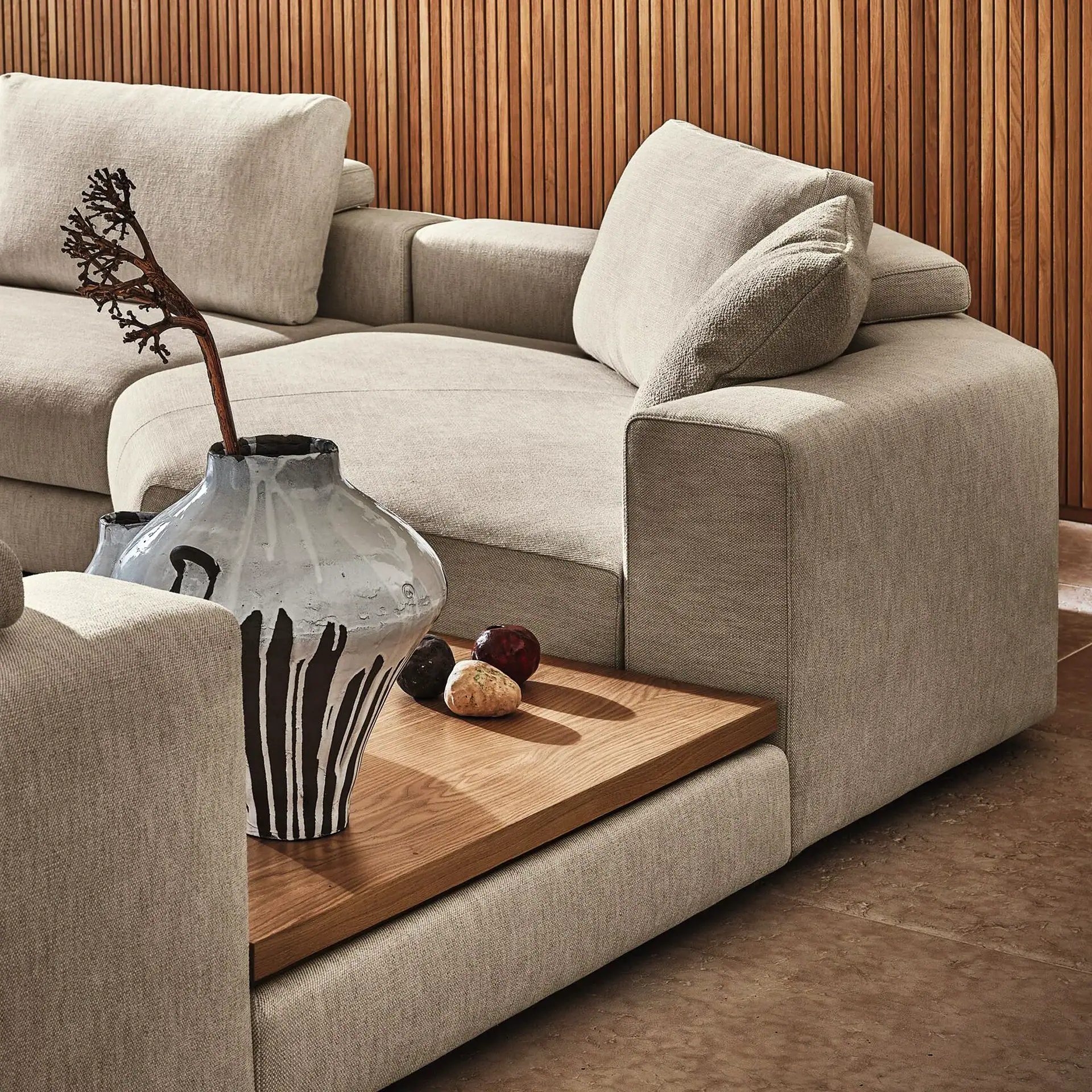 A light beige sectional sofa with an integrated wooden side table. On the table, a grey ceramic vase with black markings holds a dark branch, next to three decorative stones. A wooden slatted wall is in the background.