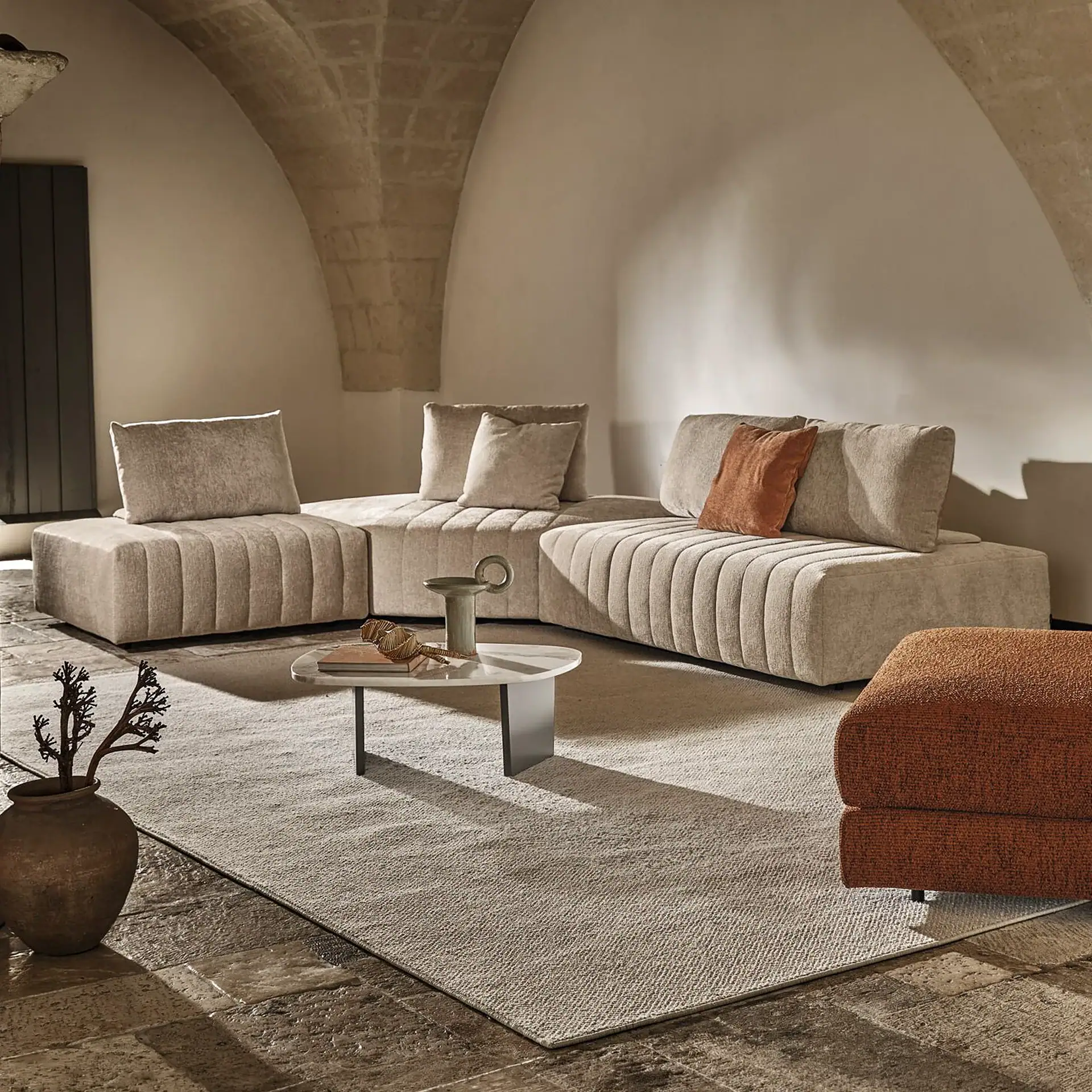 A modern living room with a modular beige ribbed sofa, a white oval coffee table, and a light-colored rug on a stone floor, under arched stone ceilings. A rust-colored cushion and ottoman add warmth.