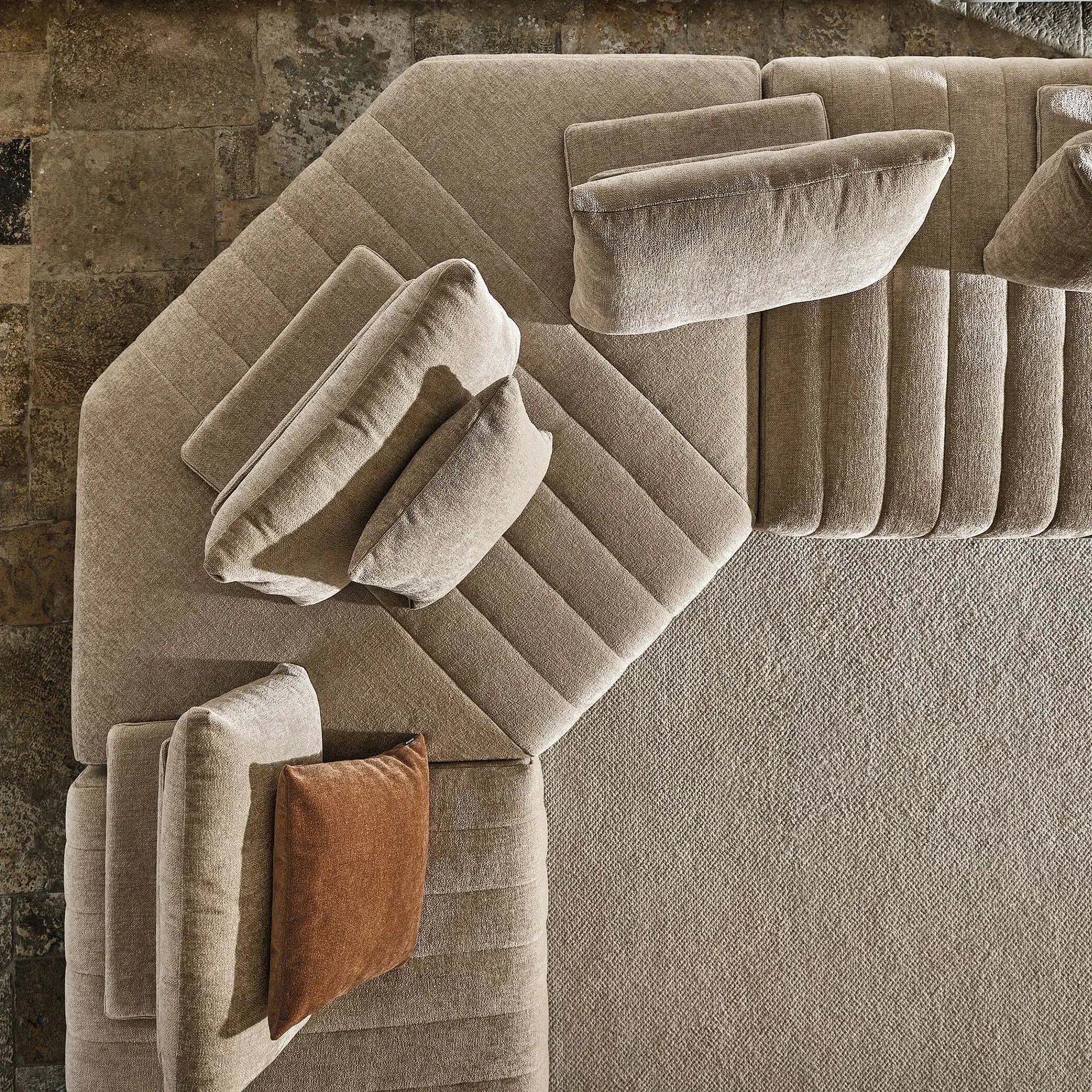 Overhead view of a large, light brown L-shaped sectional sofa with channel tufting, multiple cushions, and one burnt orange cushion, set on a textured rug and stone floor.