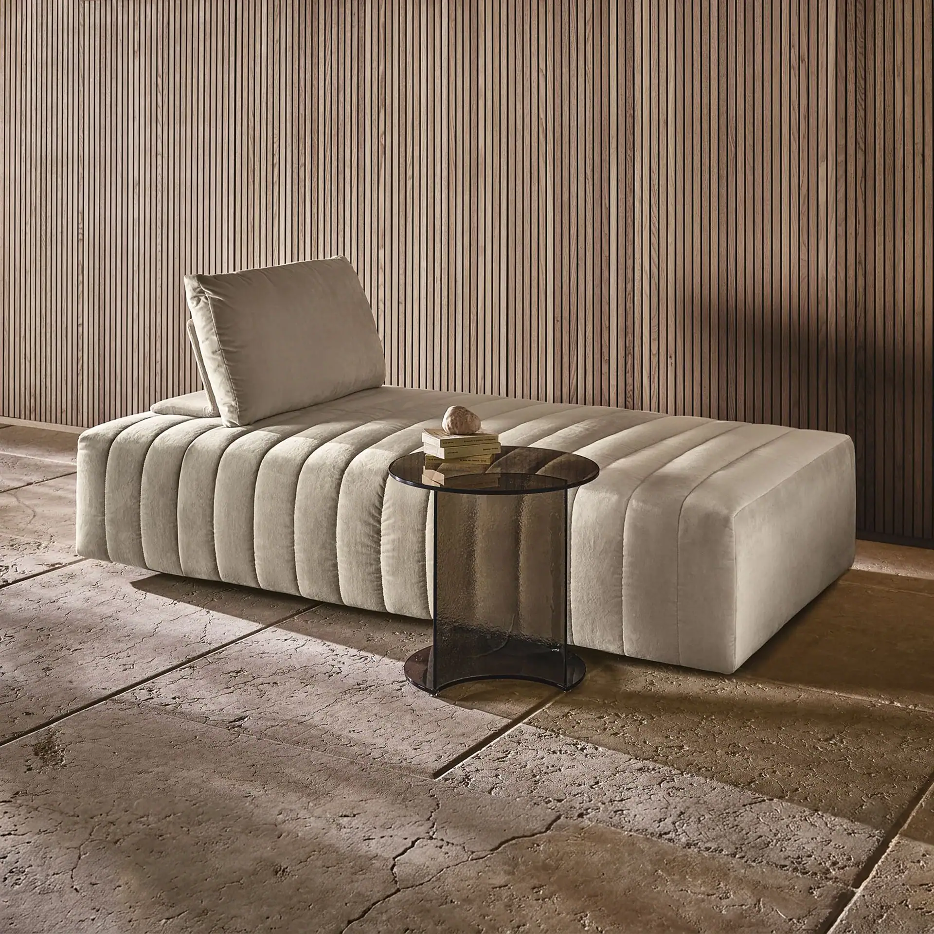 A light beige channeled velvet daybed with a matching pillow, next to a dark smoked glass side table holding books and a stone. The background is a vertical wooden slatted wall, and the floor is textured light brown stone.