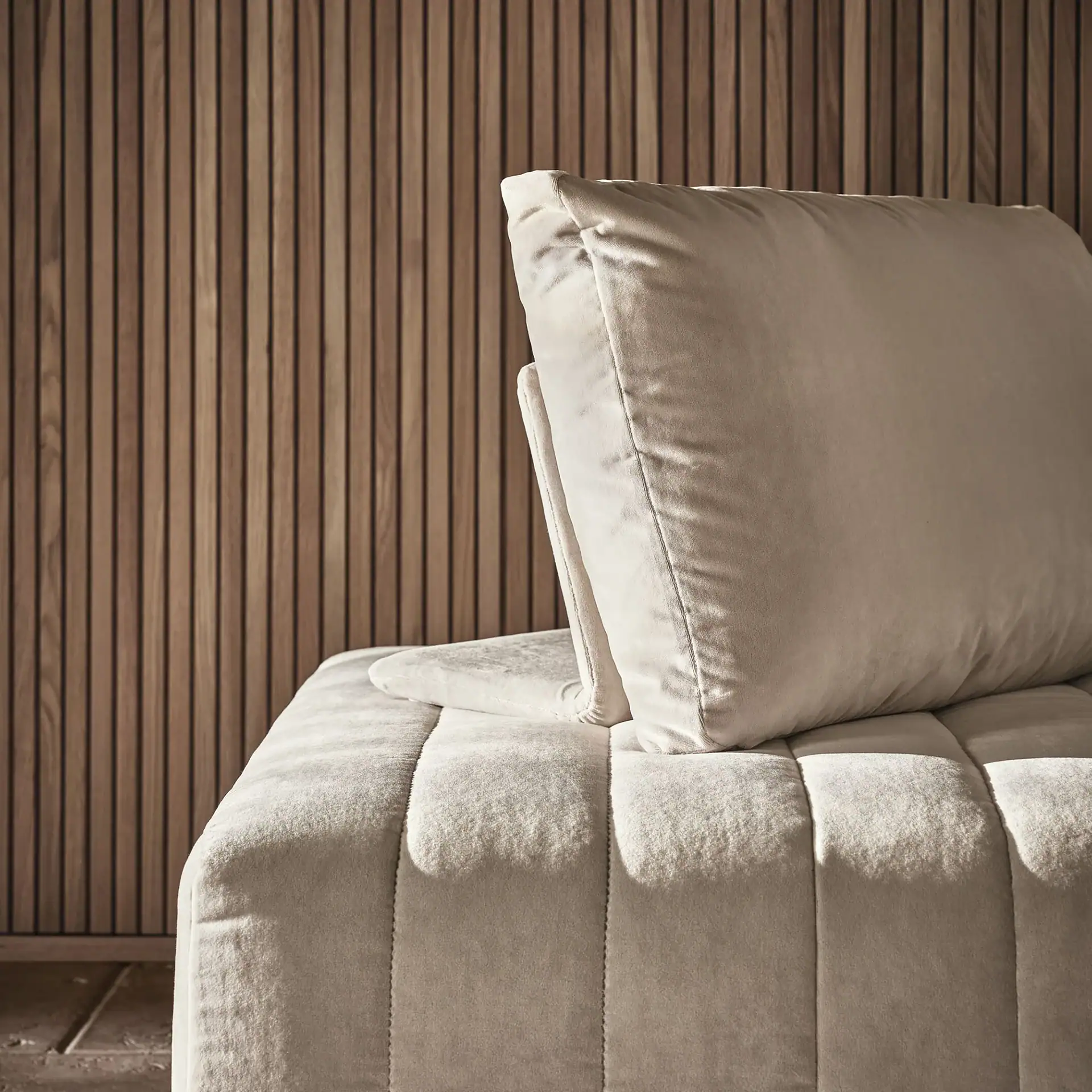 Close-up of a light beige upholstered sofa with vertical stitching, set against a warm brown wooden slatted wall.