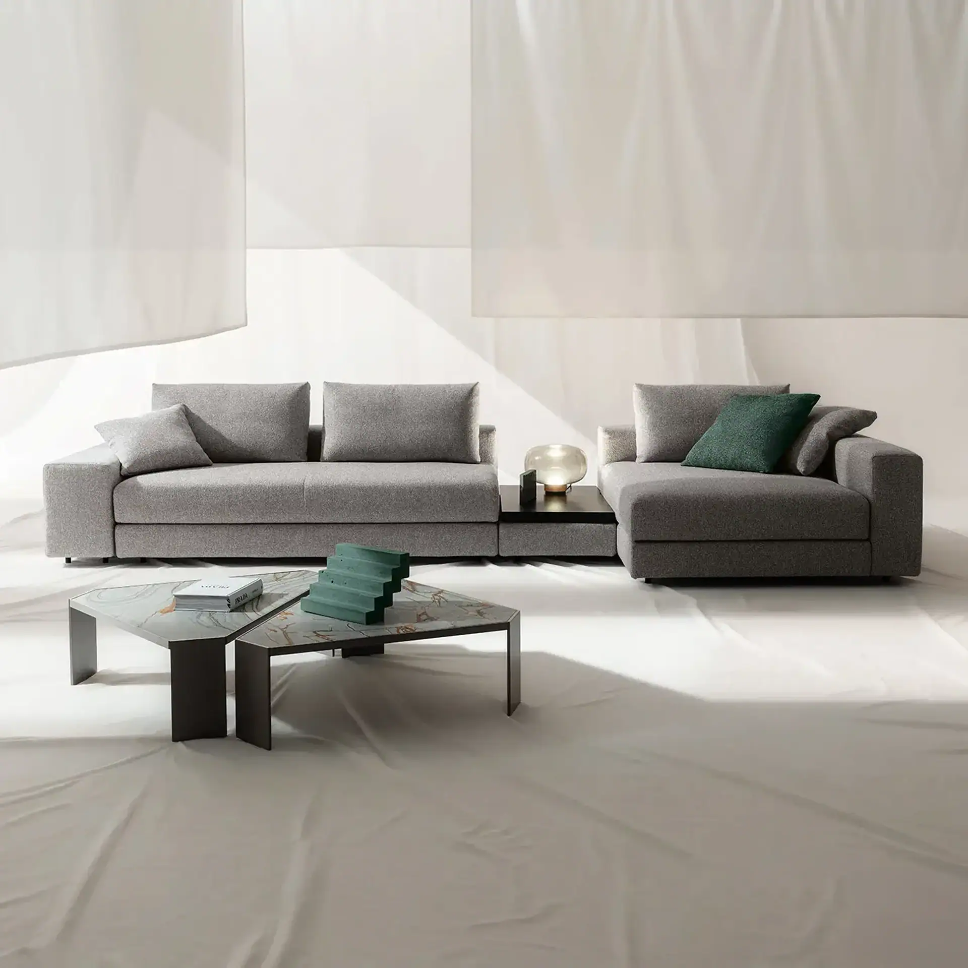 A modern grey modular sofa with a dark green throw pillow, a spherical lamp, and a marble-topped coffee table with a book and green sculpture, all against a white draped background.