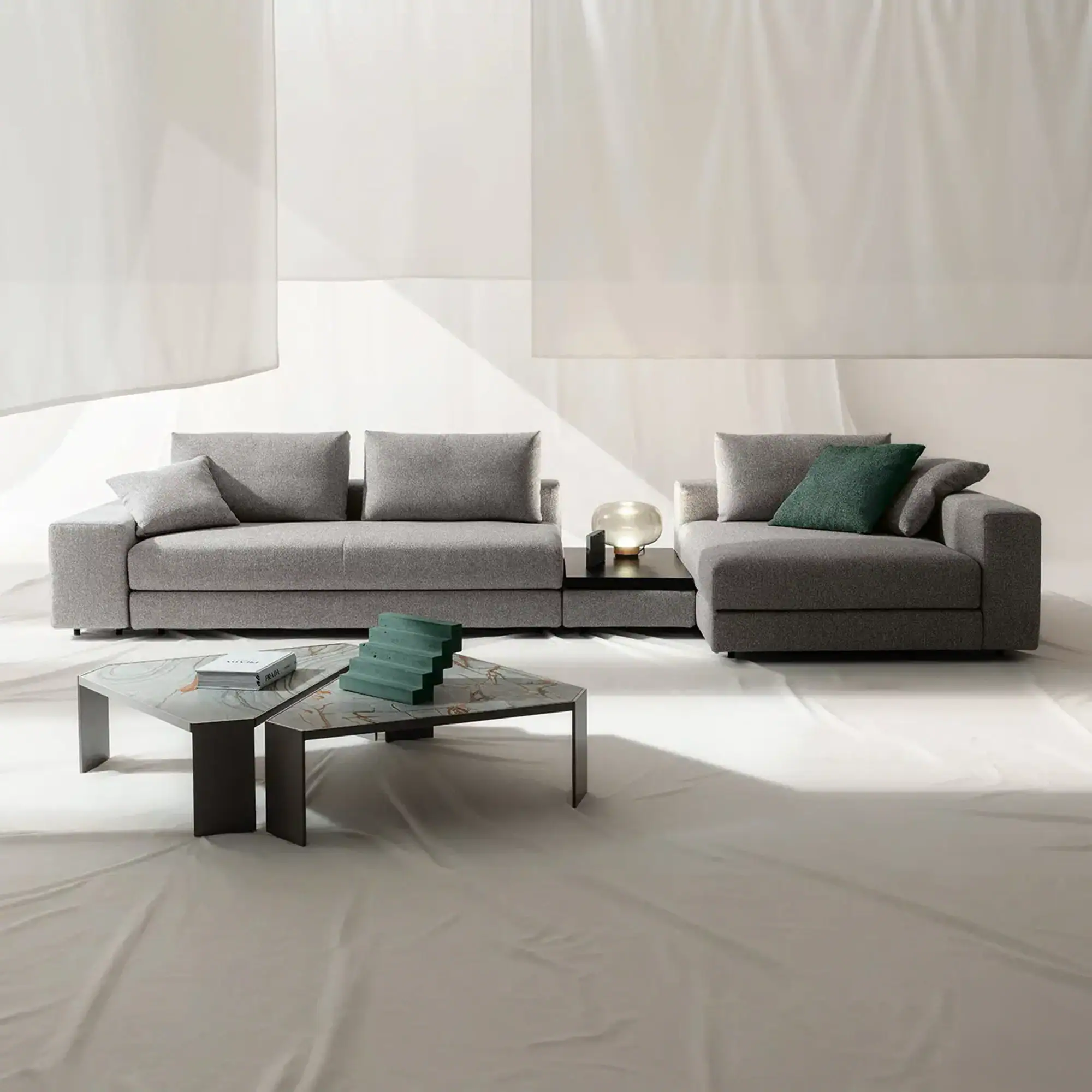 A modern grey modular sofa with a dark green throw pillow, a spherical lamp, and a marble-topped coffee table with a book and green sculpture, all against a white draped background.