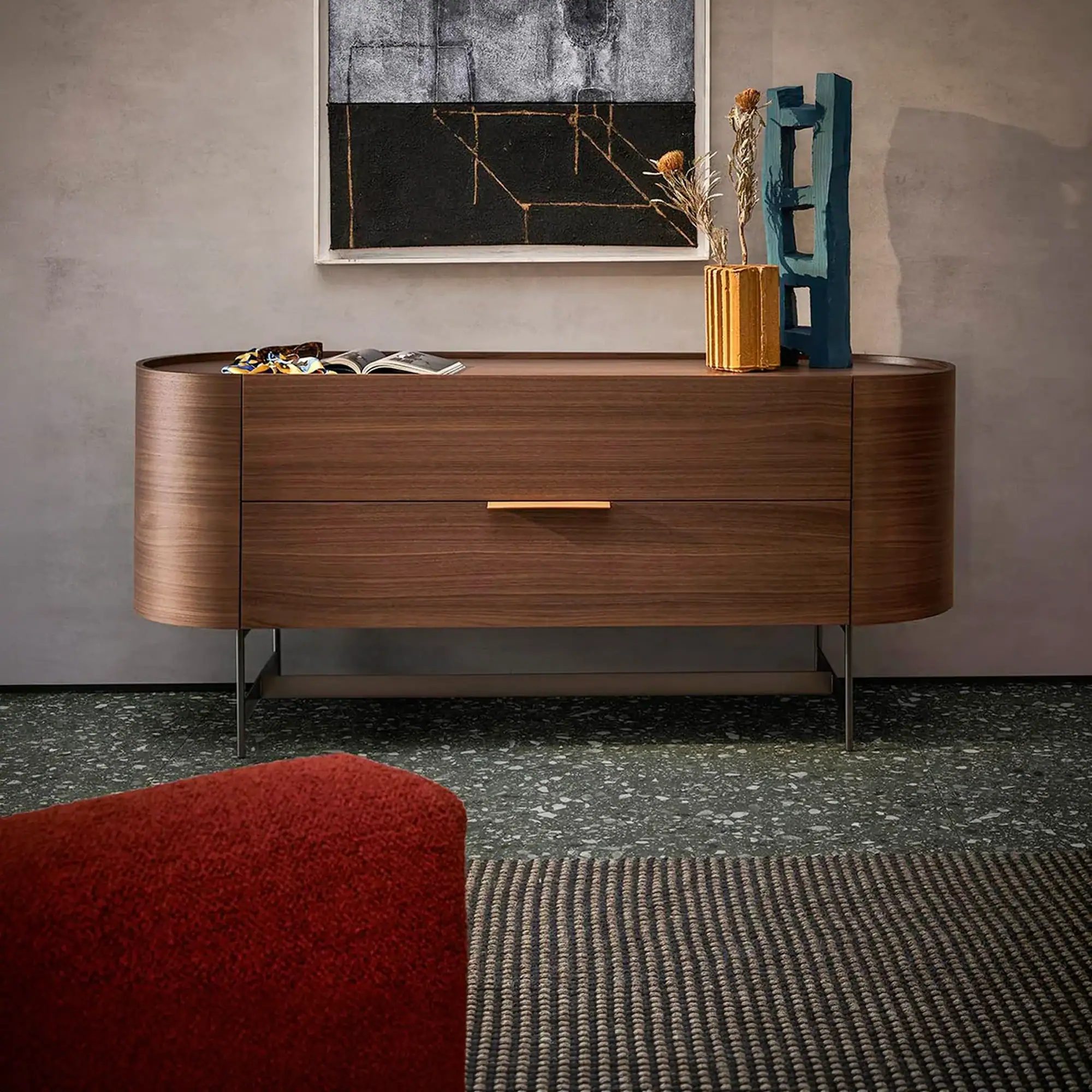 A modern dark wood sideboard with rounded edges, featuring an abstract painting above and decorative items on top, including a golden vase and a teal sculpture. A red armchair and a striped rug are partially visible on a terrazzo floor.
