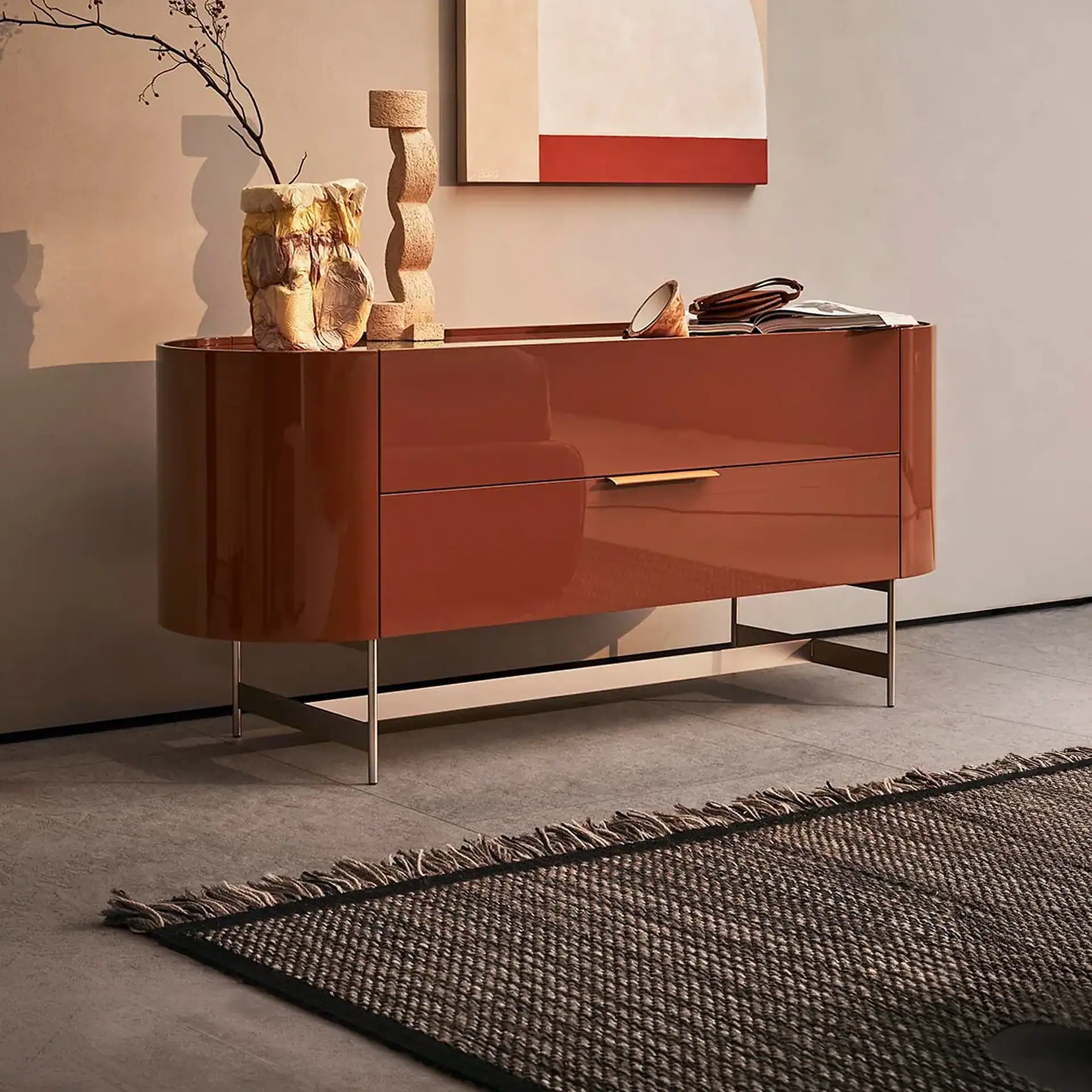 A modern, glossy reddish-brown dresser with gold handles and metal legs, topped with a textured vase holding a branch, a sculptural cork object, a small bowl, an open book, and a wallet. A minimalist painting hangs above, and a woven rug is on the tiled floor.