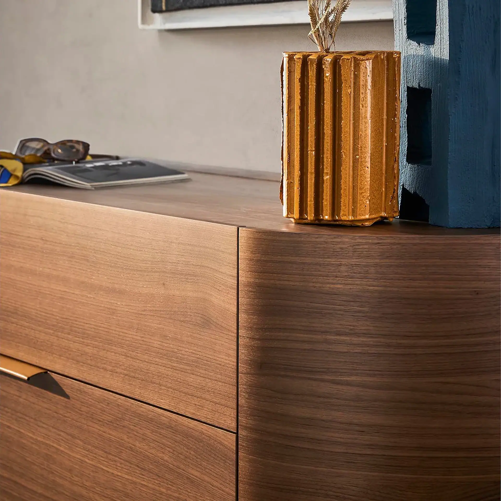 Close-up of a dark brown wooden dresser with a rounded corner. On top are sunglasses, a magazine, a golden ribbed vase with dried grass, and a blue textured block.