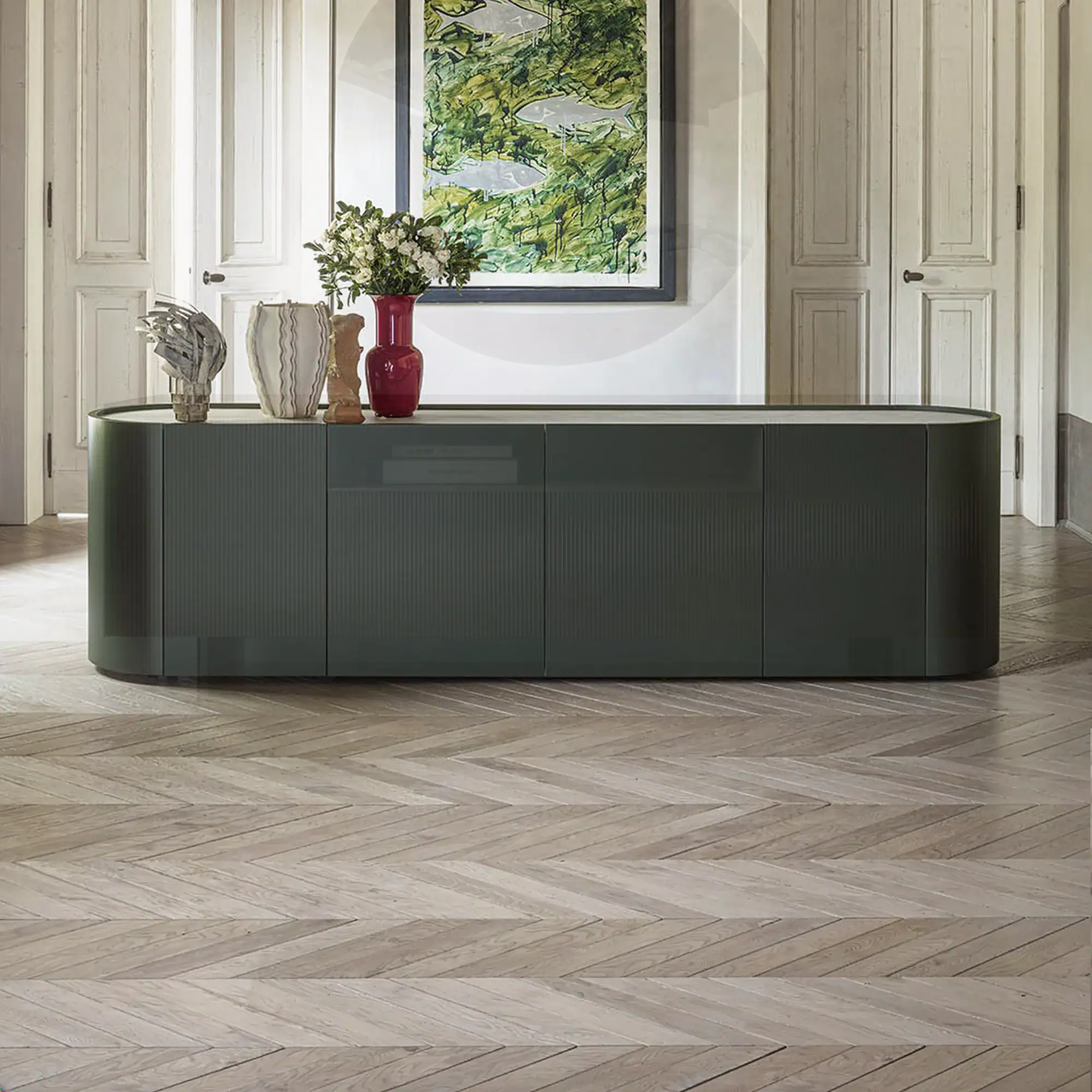 A dark green sideboard with rounded edges, decorated with vases and sculptures, sits on a herringbone wood floor in front of white paneled doors and an abstract green painting.