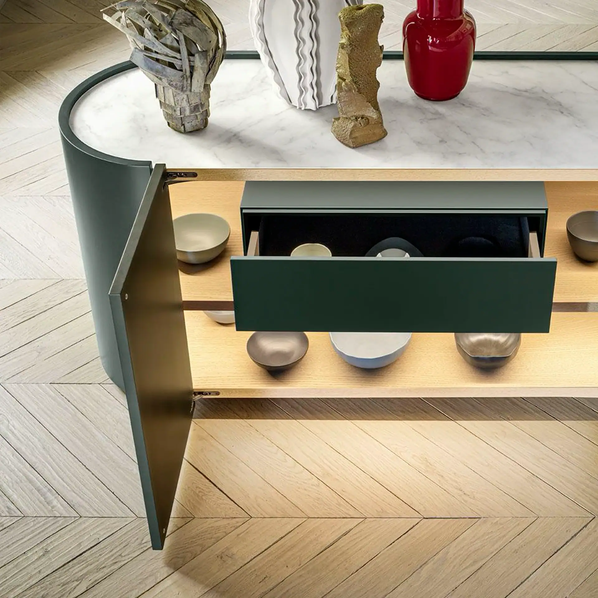 A dark green console table with a white marble top, open drawer, and decorative items on a light herringbone wooden floor.