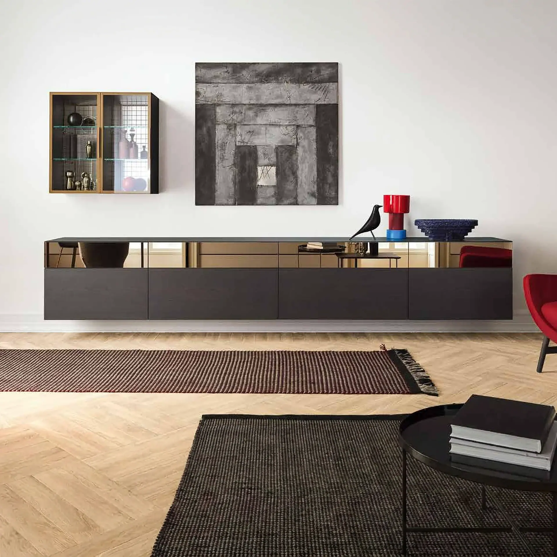 A modern living room with a dark floating console, abstract grey painting, wall-mounted display cabinet, and herringbone wood floor with two striped rugs.