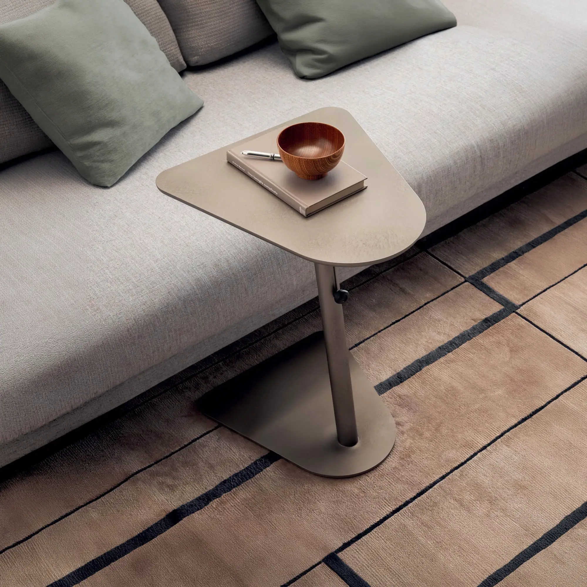 A modern side table with a book and wooden bowl next to a light-colored sofa with green pillows, on a geometric patterned rug.