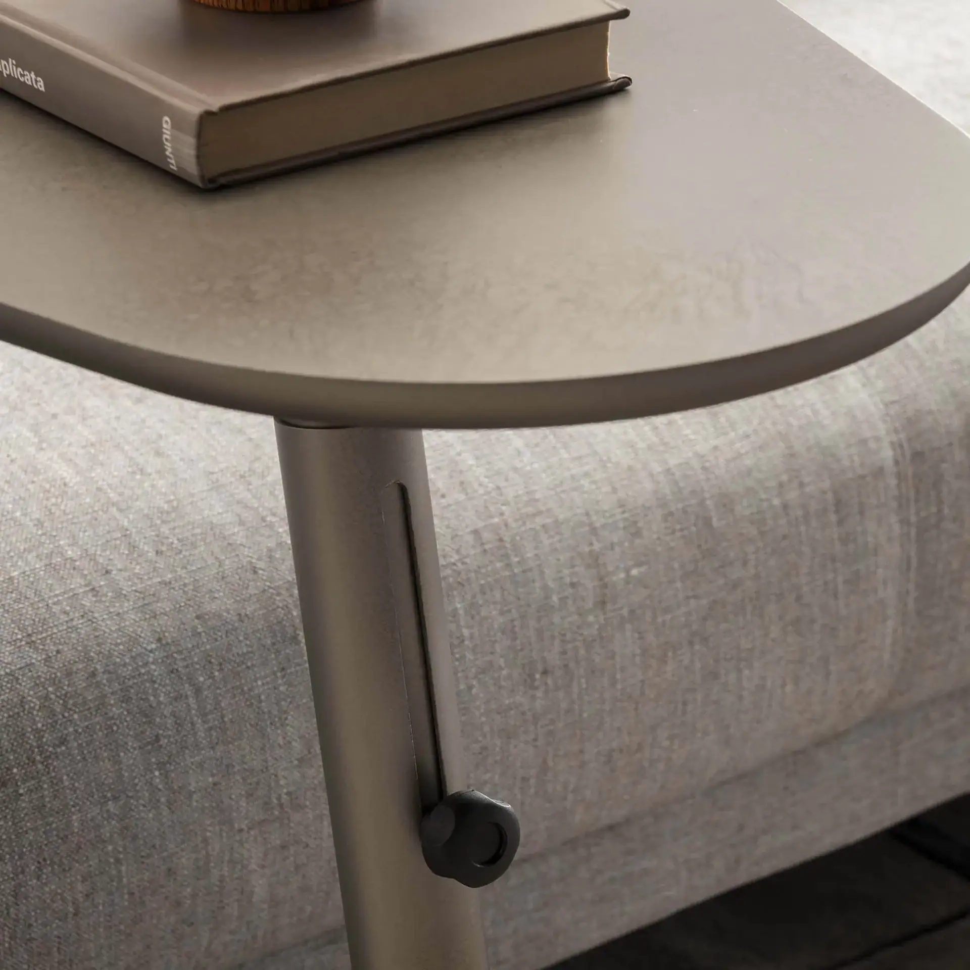A close-up of a modern, matte grey-taupe adjustable side table with a book on top, next to a textured, light grey upholstered sofa.