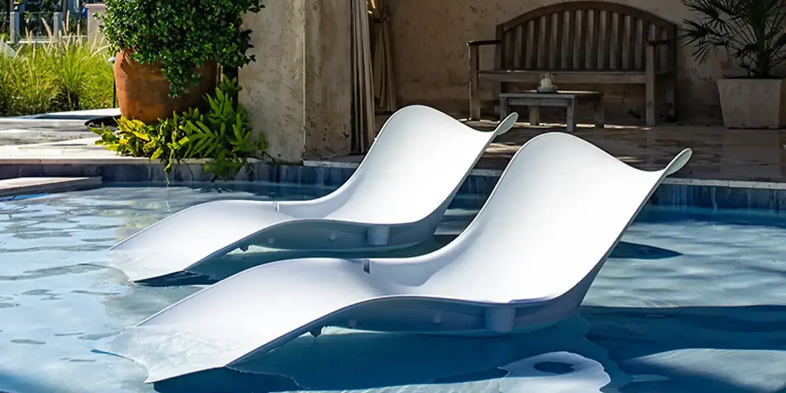 Two white, curved lounge chairs are partially submerged in a light blue swimming pool, with a patio, wooden bench, and green plants in the background. Ocean In Pool Chaise Lounge by Couture Jardin