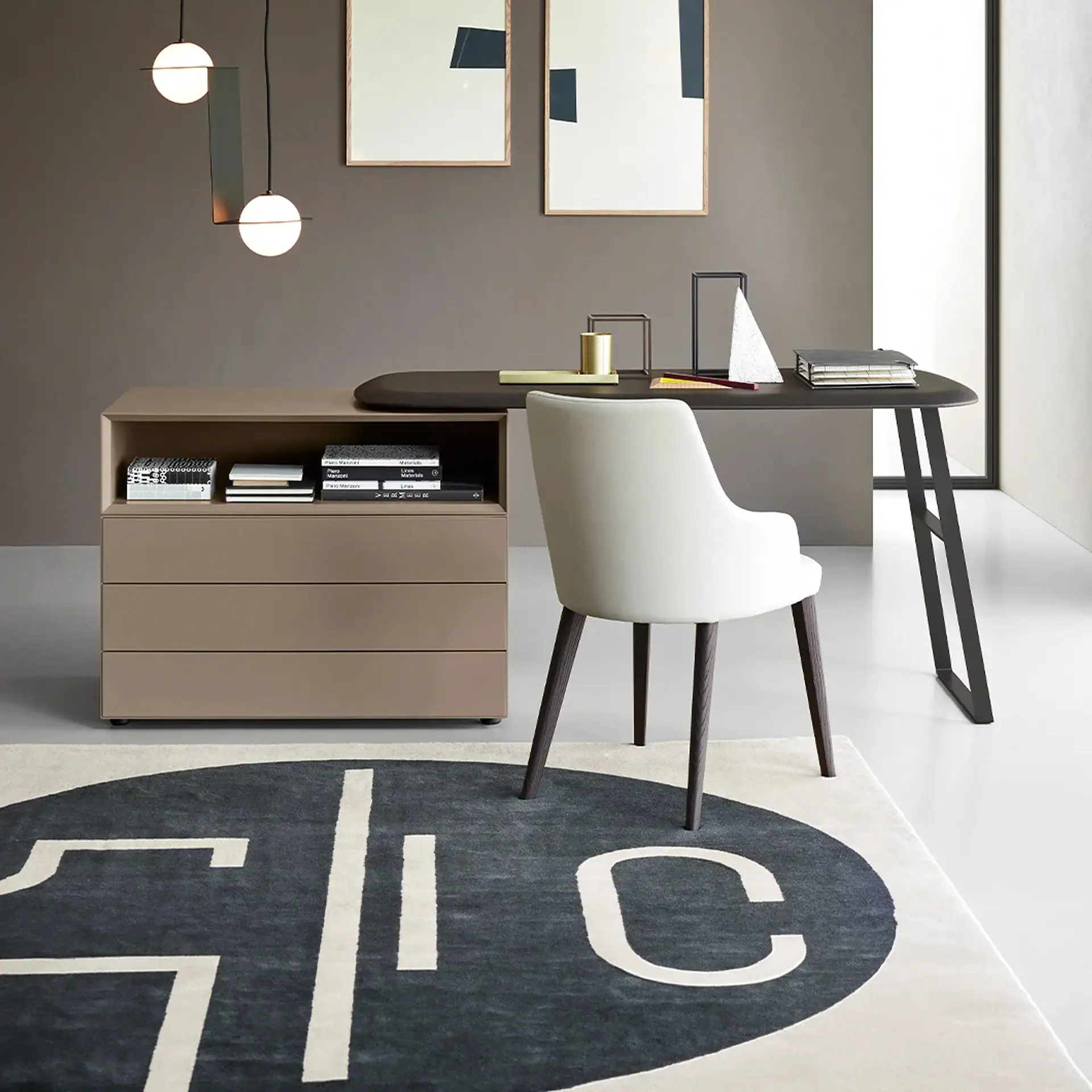 A modern home office with a dark oval desk, cream chair, taupe storage unit, and abstract art on a dark taupe wall.