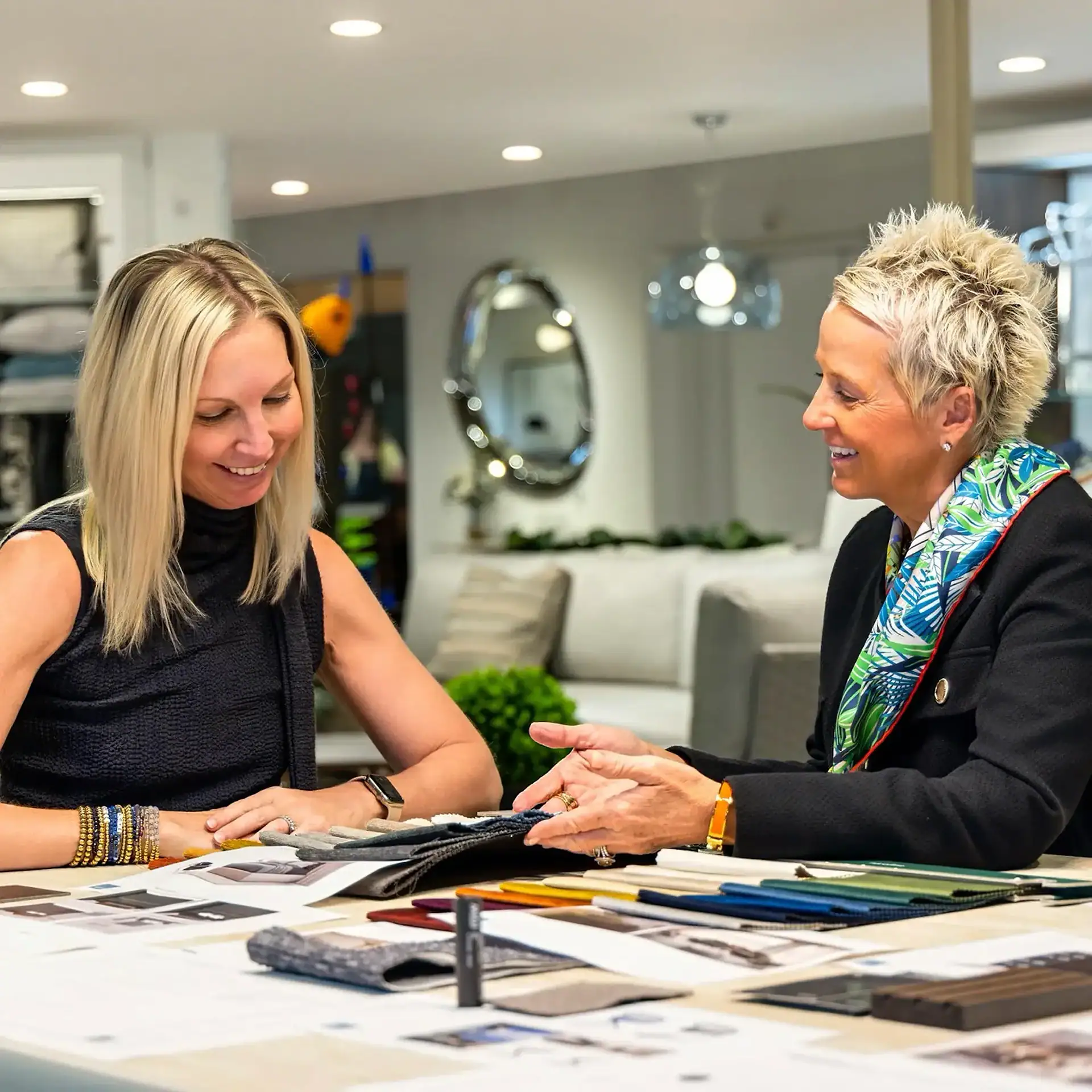 Two women, one with long blonde hair and one with short spiky blonde hair, are seated at a table, smiling and looking at various fabric swatches and design materials spread before them.