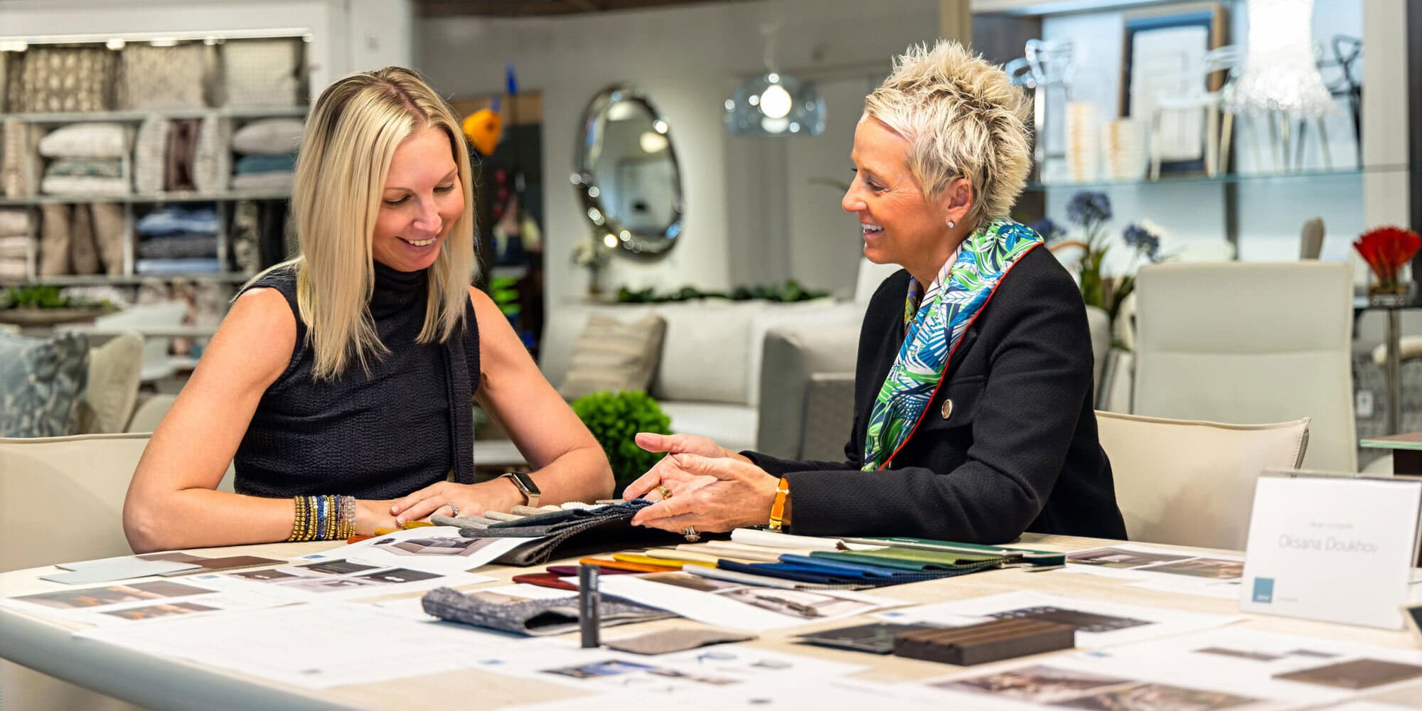 Two women smiling and discussing fabric samples at a table in a design showroom.