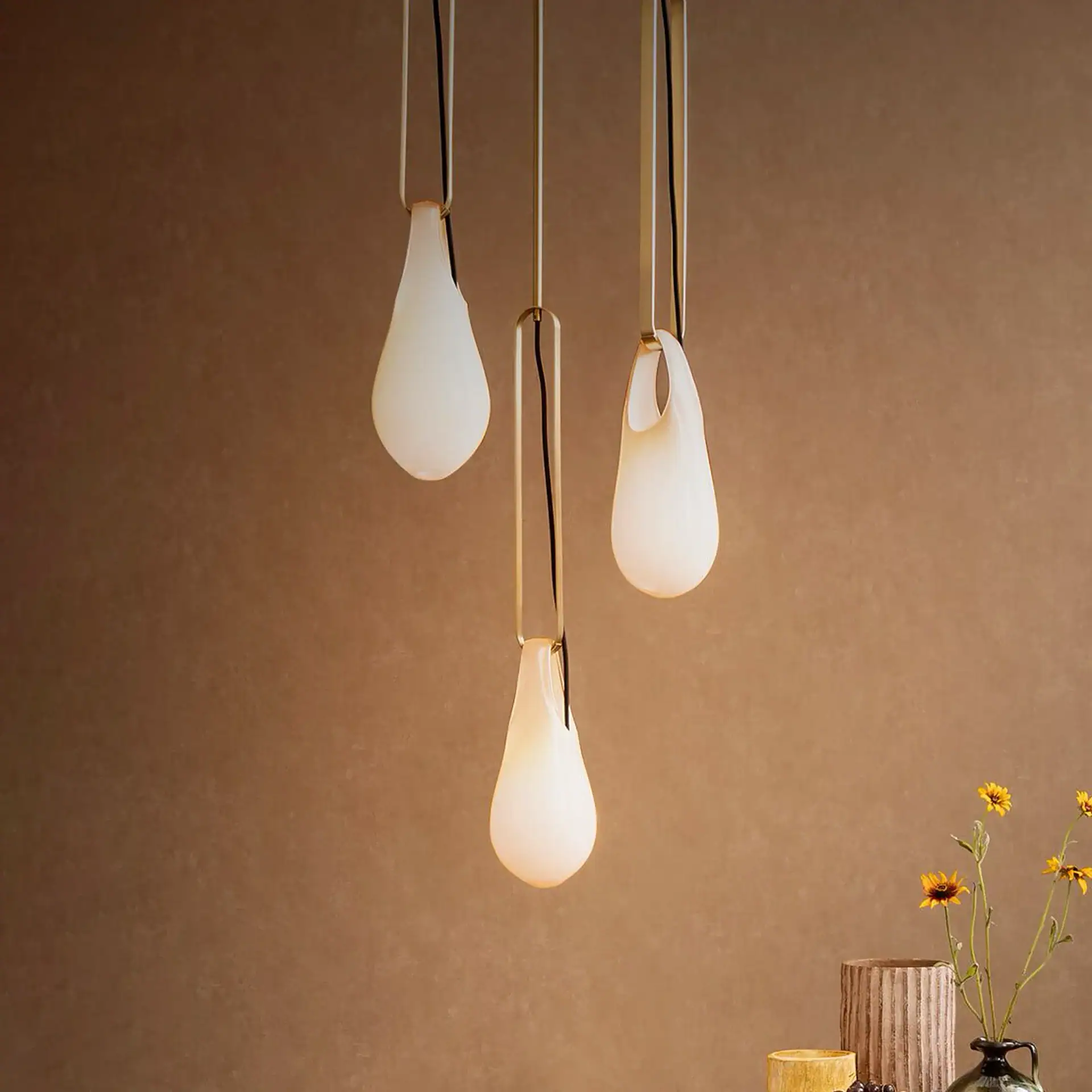 Three illuminated, organic-shaped frosted white glass pendant lights hanging against a brown wall, with a vase of yellow flowers and other decor visible in the bottom right.