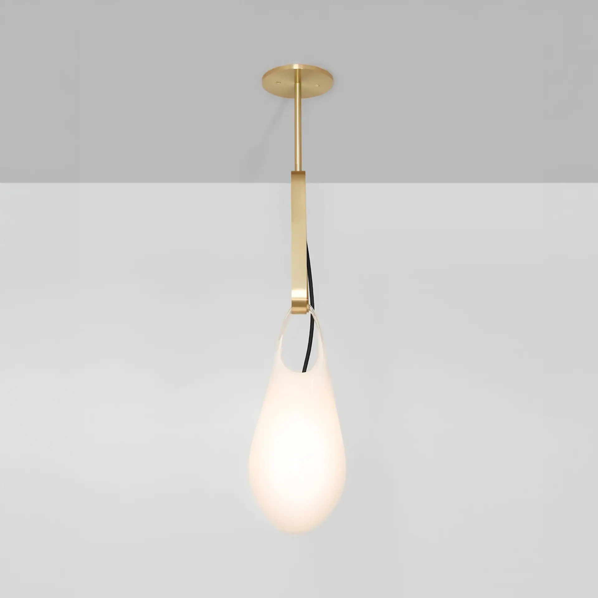 A modern pendant light featuring a brass fixture and a glowing, teardrop-shaped frosted white glass diffuser, suspended from a gray ceiling.
