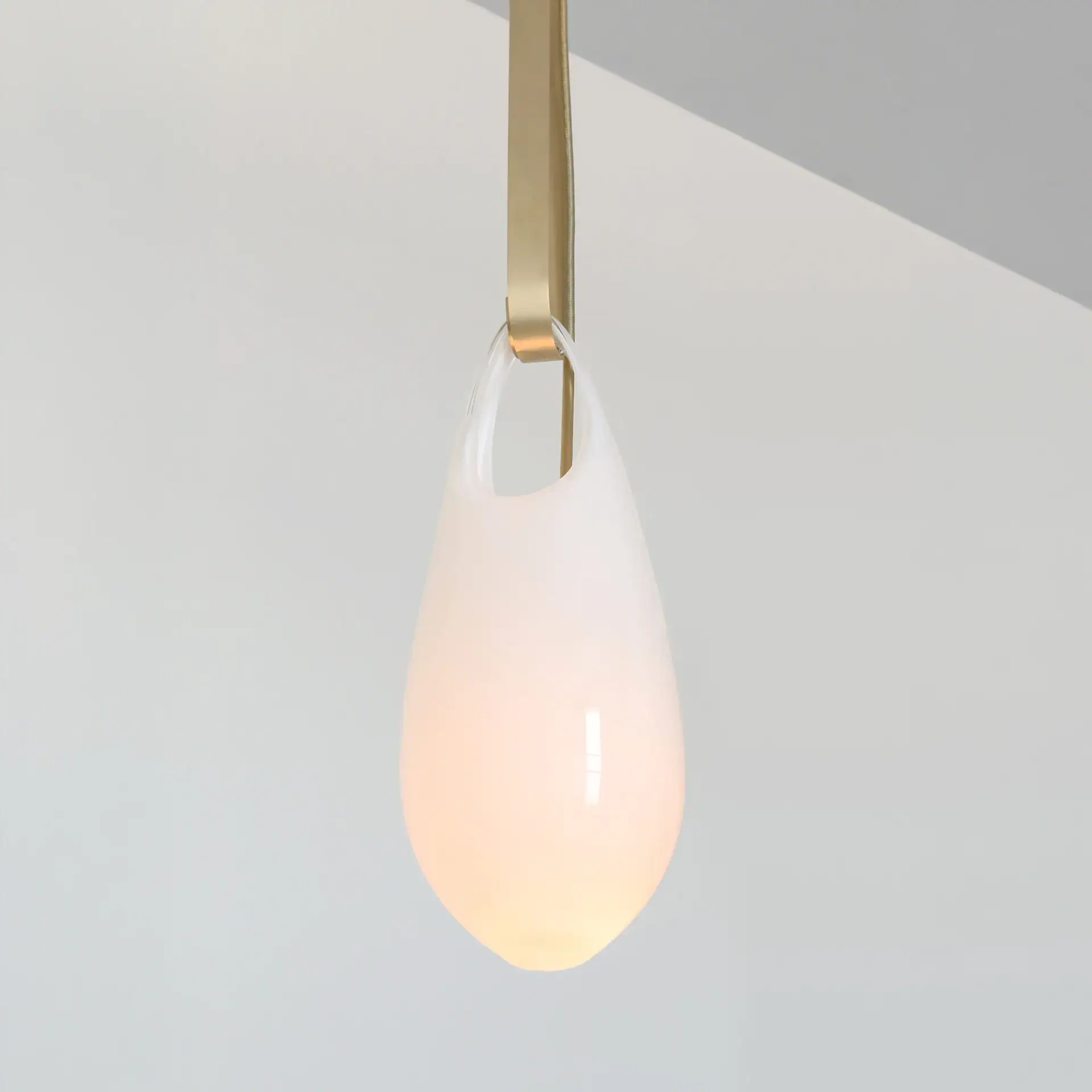A minimalist teardrop-shaped pendant light with a frosted white glass shade, glowing warmly, suspended by a brass strap against a light gray wall.