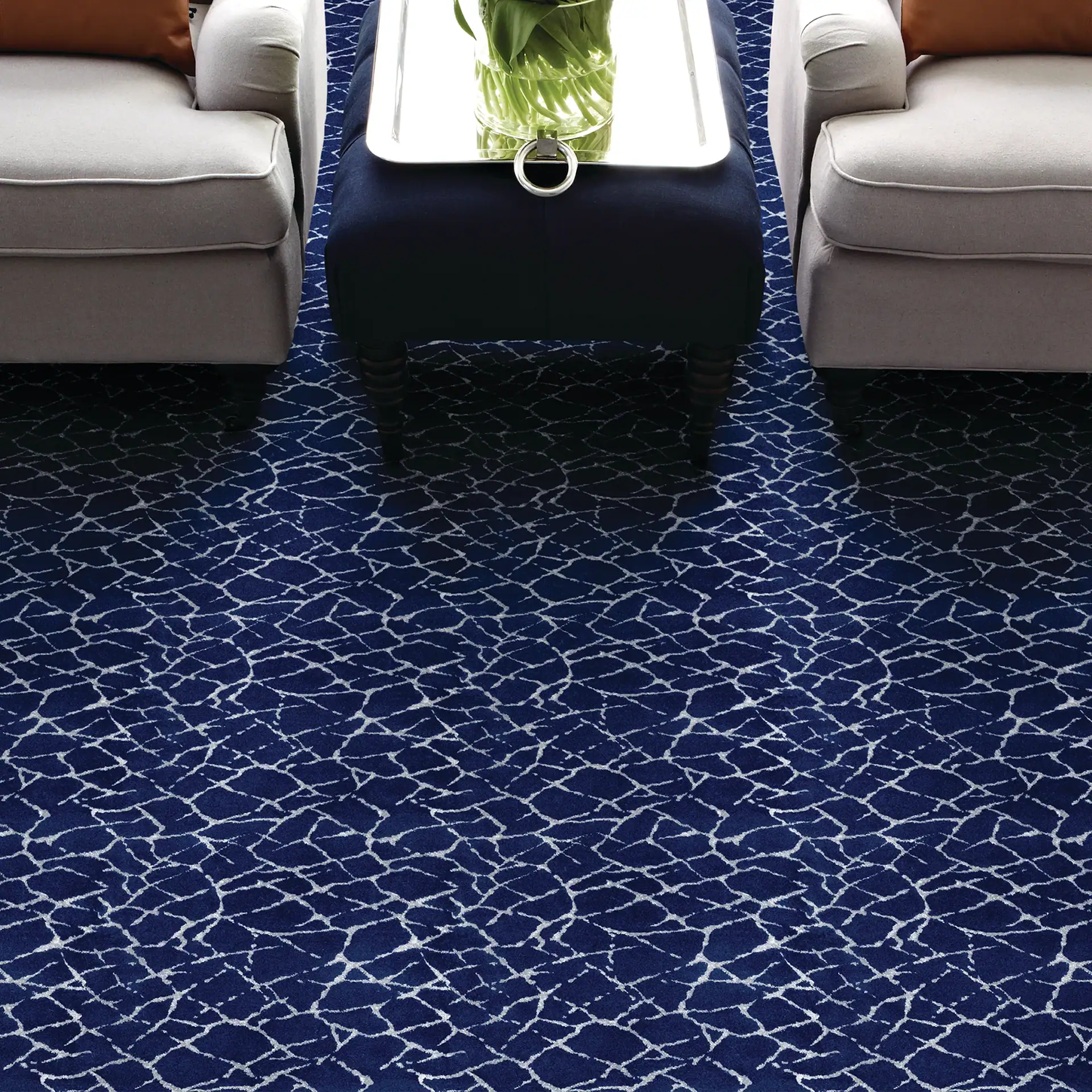 A room featuring a dark blue carpet with a white irregular pattern, two light-colored armchairs with orange pillows, and a blue ottoman with a silver tray and a glass vase of green stems.