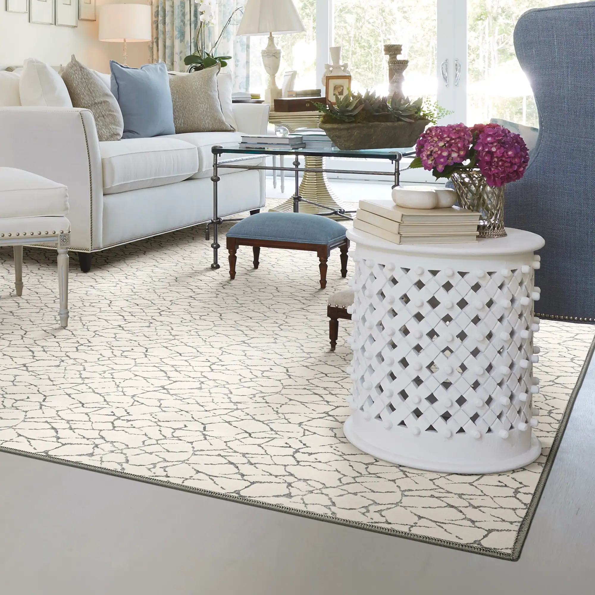 A brightly lit living room with a white sofa, blue wingback chair, glass coffee table, and a cream rug with a grey pattern.