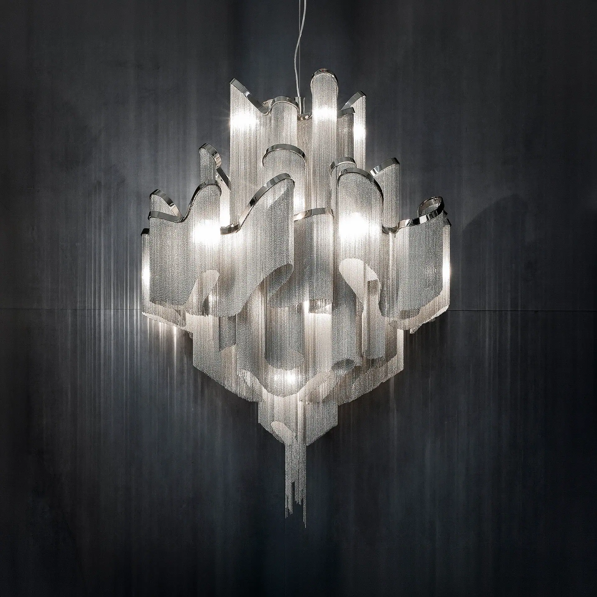 A large, modern chandelier made of shimmering silver metal chains and polished frames, brightly lit against a dark background.