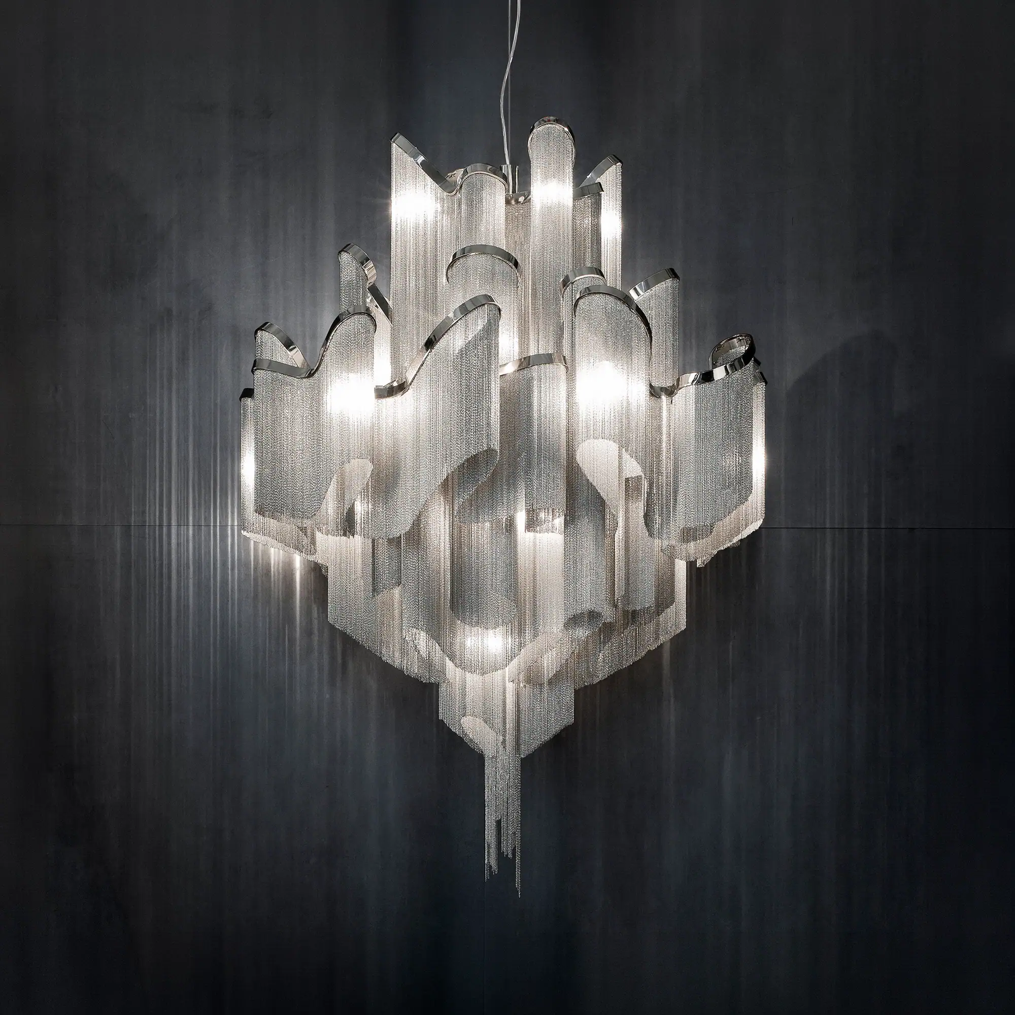 A large, modern chandelier made of shimmering silver metal chains and polished frames, brightly lit against a dark background.