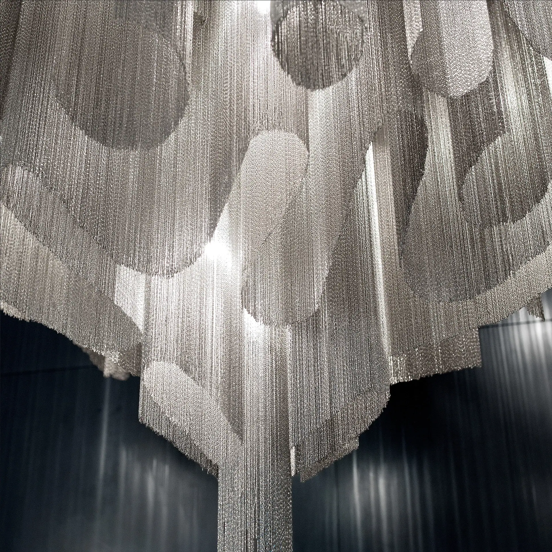 A large chandelier made of shimmering silver chains forming intricate circular and wavy patterns, illuminated from within against a dark background.