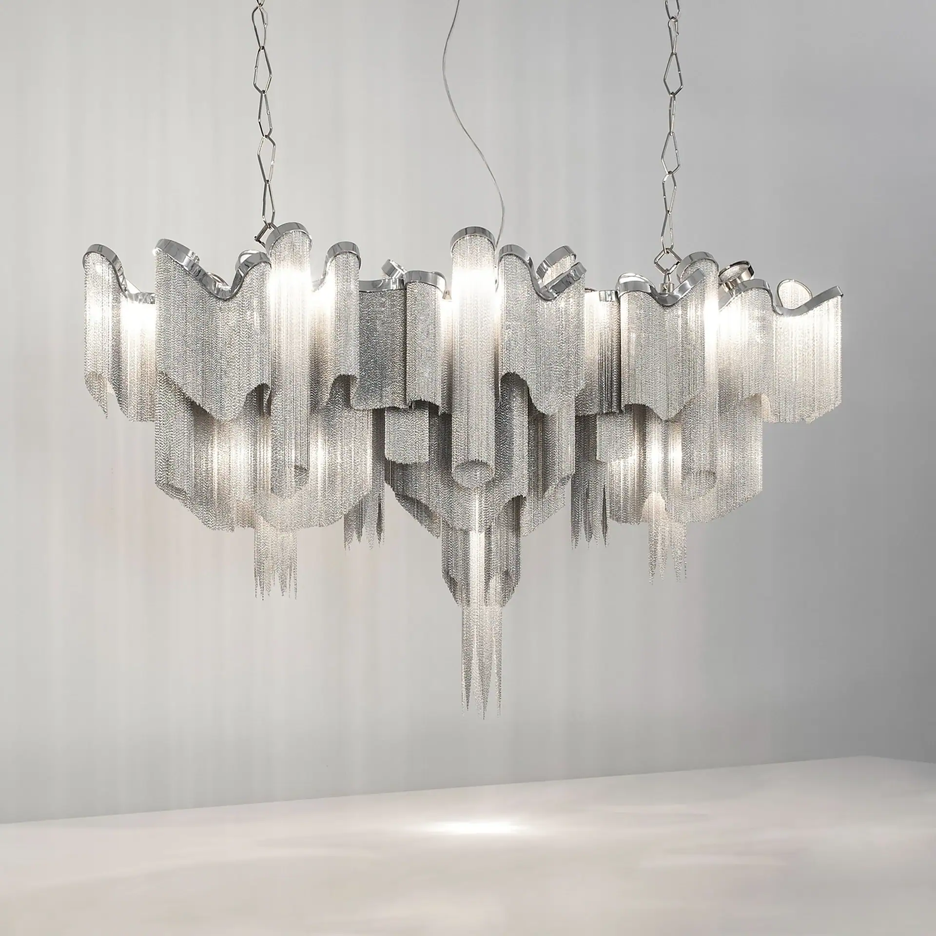 A modern chandelier made of cascading silver metal chains, illuminated from within, hanging against a light gray background.