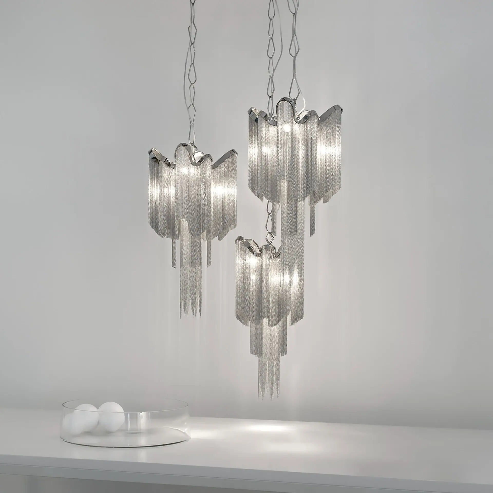 Three modern pendant chandeliers made of cascading silver chains hang against a light gray wall, with a glass bowl containing two white spheres on a white surface below.