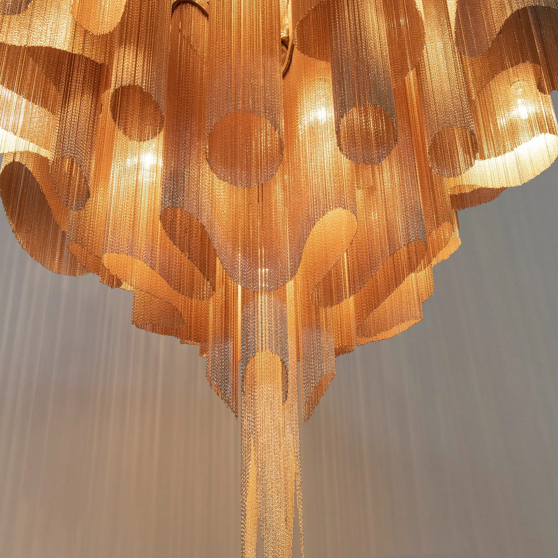 A large, ornate chandelier made of numerous strands of golden and silver metallic chains, illuminated from within with a warm light.