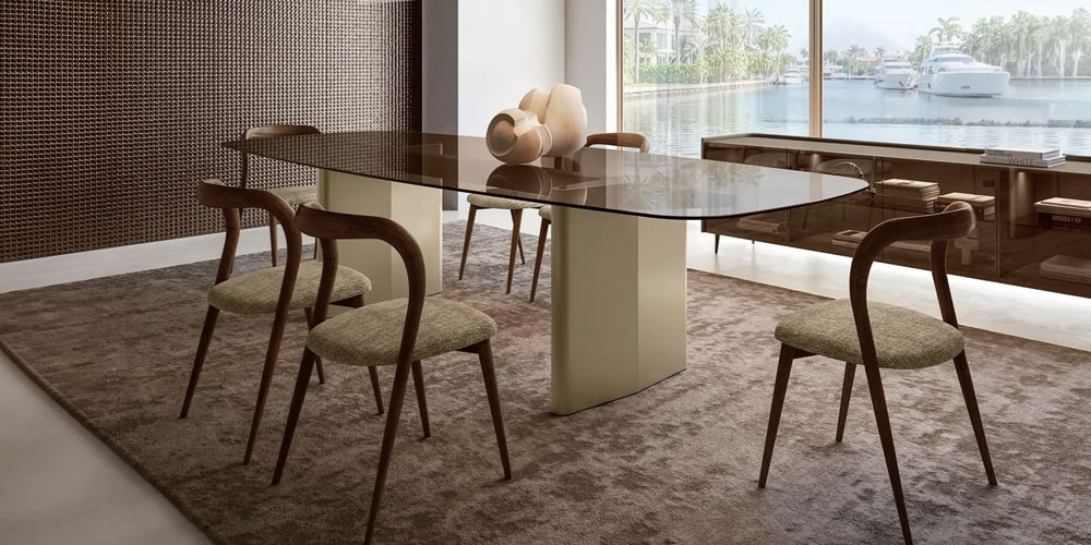 A modern dining room with a glass table, six wooden chairs, and a large window overlooking a waterfront with yachts.