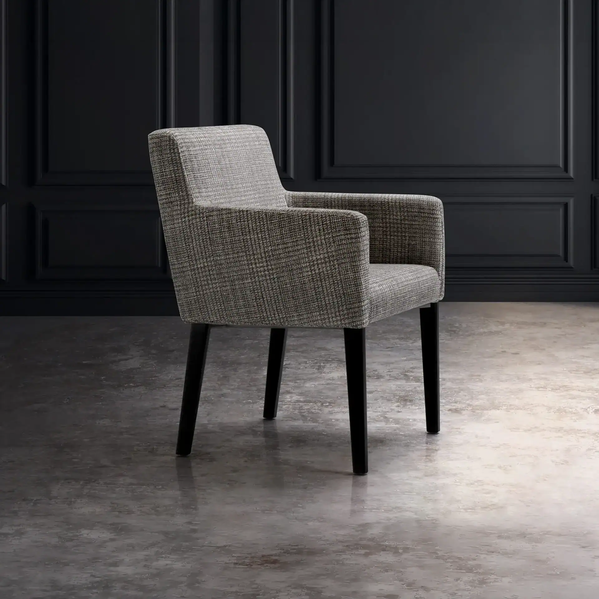 A modern gray speckled upholstered armchair with dark legs sits on a concrete floor in front of a dark gray paneled wall.