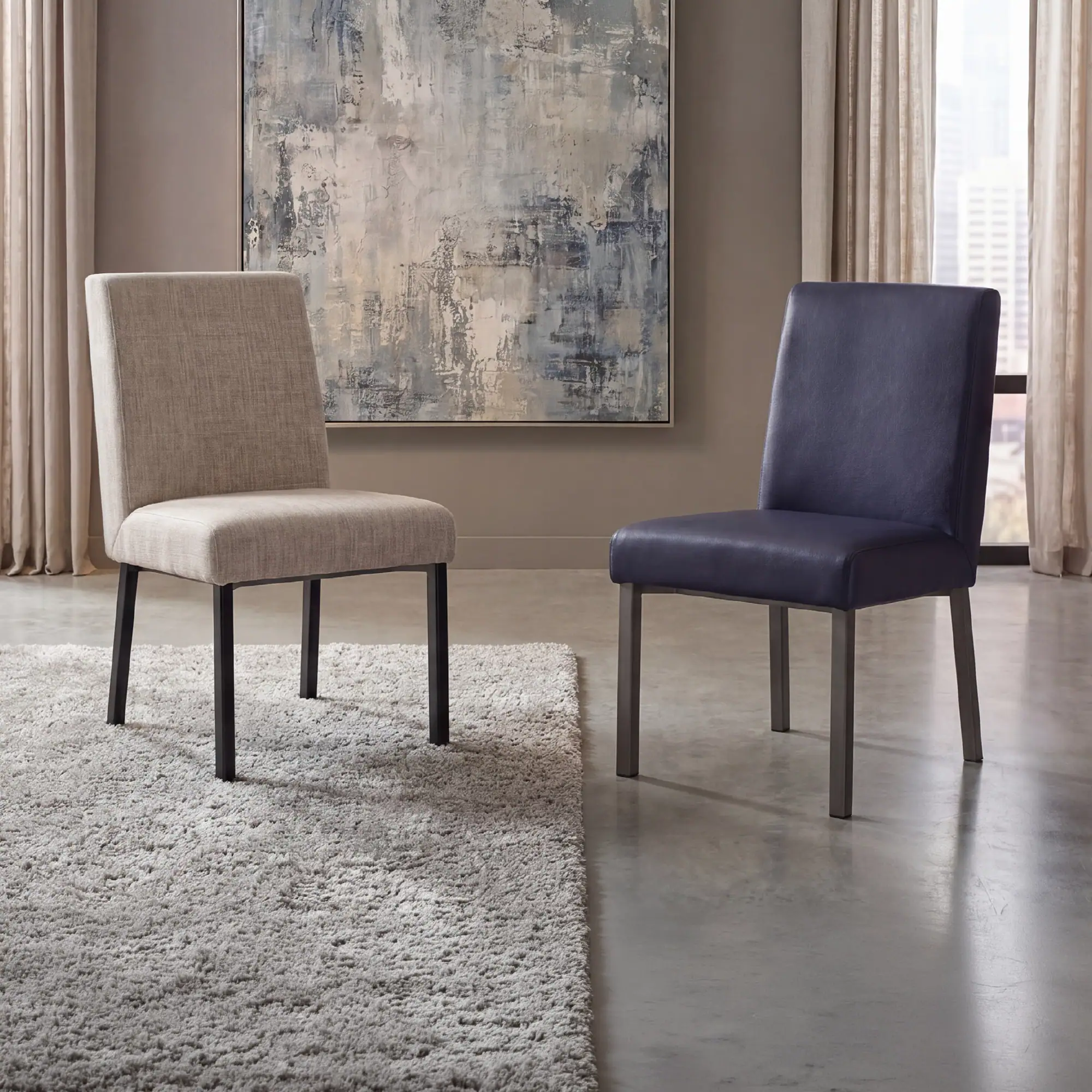 Two modern chairs, one beige fabric with black legs and one navy leather with silver legs, in a room with an abstract painting and a city view.
