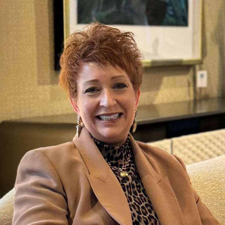 A smiling woman with short reddish-brown hair wearing a leopard print top and brown blazer.
