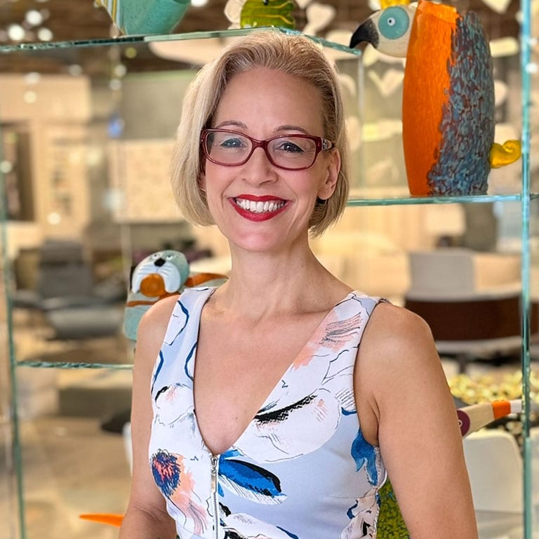 A smiling woman with short blonde hair and red glasses, wearing a floral dress, stands in front of glass shelves with decorative art.