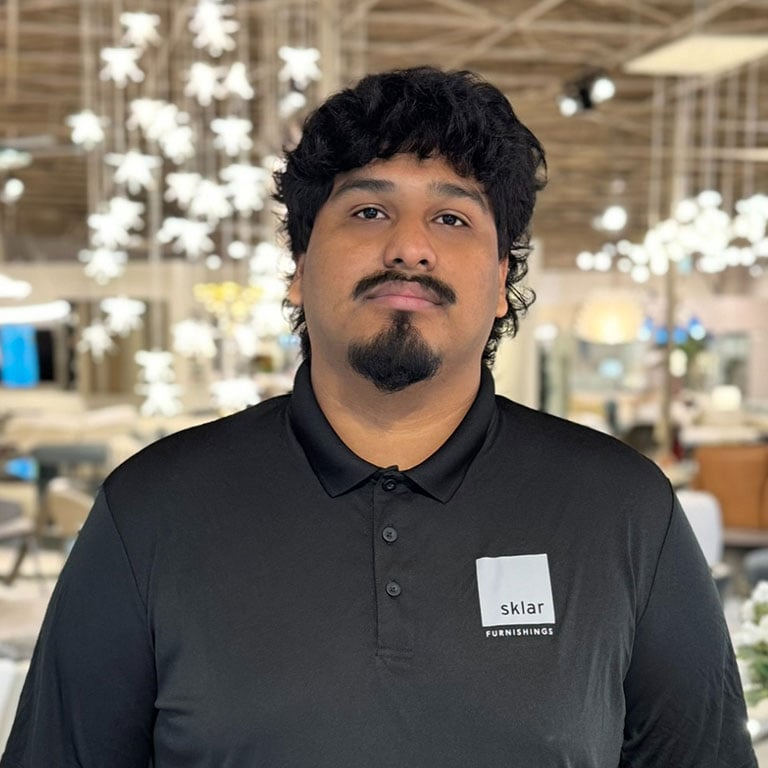 A man with dark hair and a goatee wearing a black polo shirt with the Sklar Furnishings logo. He looks directly at the viewer in a blurred showroom background.