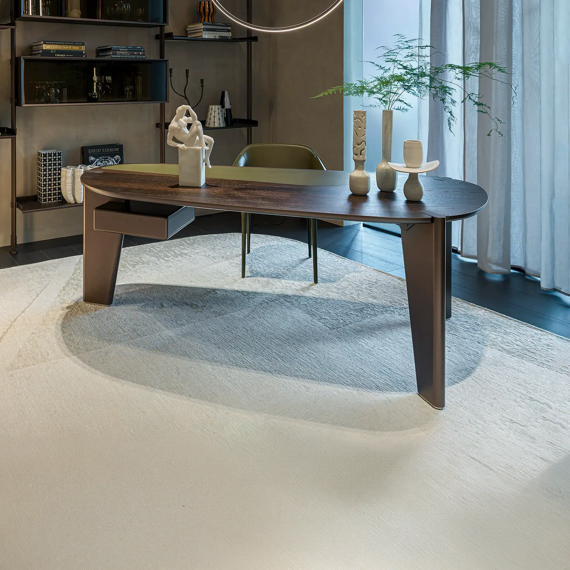A modern home office with a dark wood oval desk, green leather inlay, white abstract sculpture, and fern plant. A dark metal bookshelf and sheer white curtains are in the background, with a light-colored abstract rug on the floor.