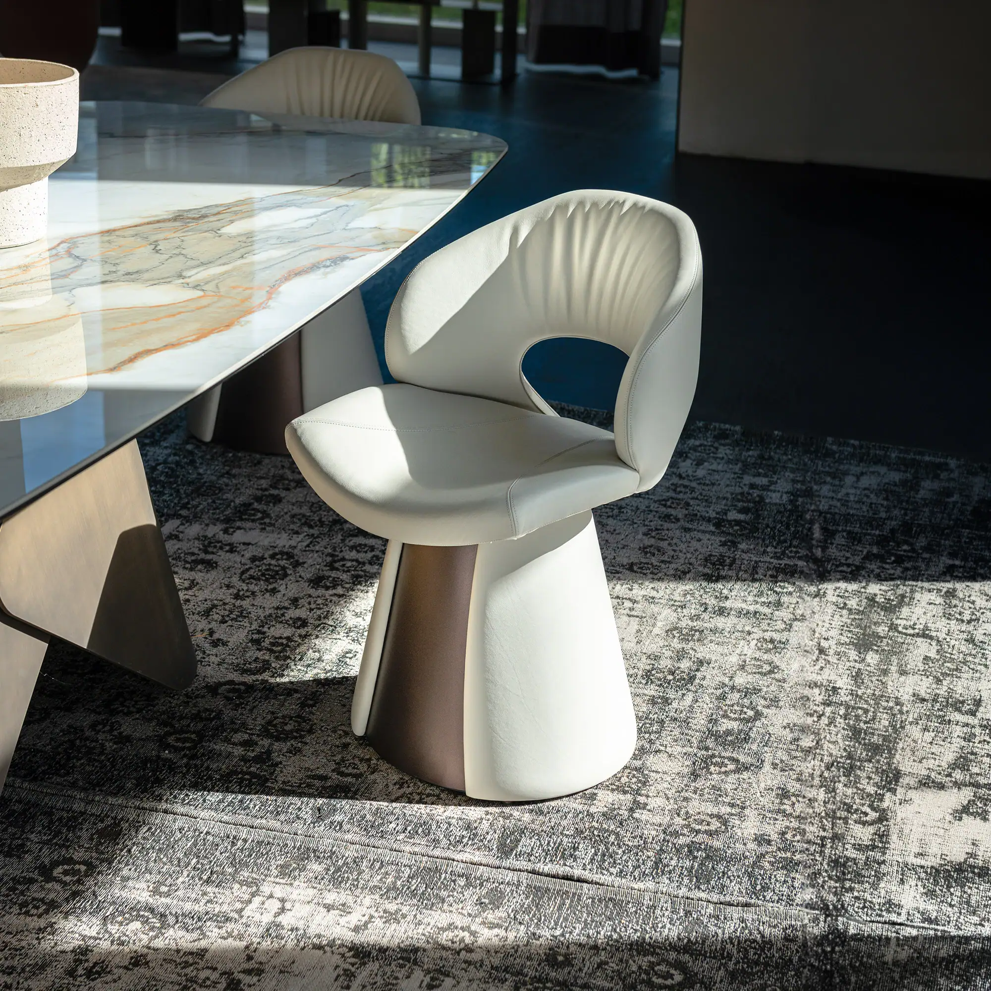 A cream leather modern dining chair with a two-toned conical base, next to a marble-top dining table with a textured vase, all on a patterned rug.