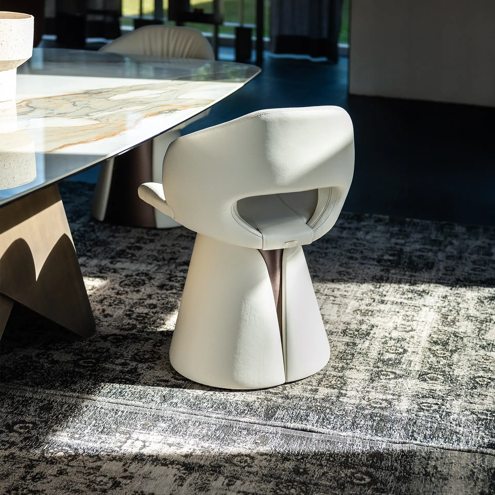 A modern cream leather dining chair with a unique cut-out back and conical base, next to a marble-patterned dining table on a patterned rug, illuminated by natural light.