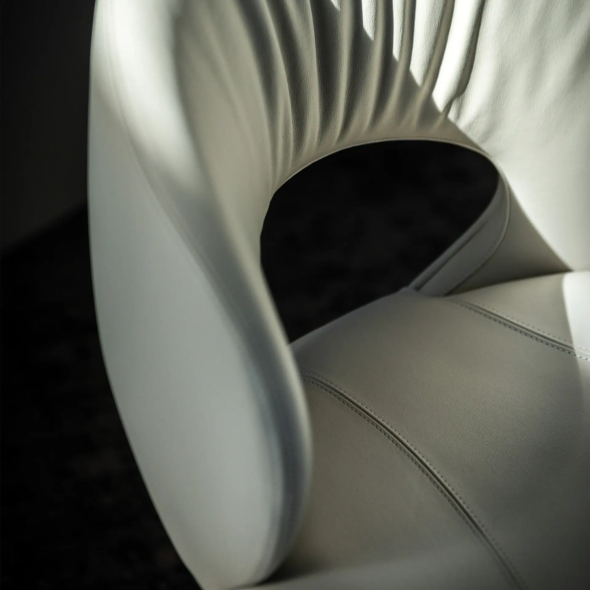 Close-up of a white leather chair, showing pleated curves, stitching, and strong shadows against a dark background.