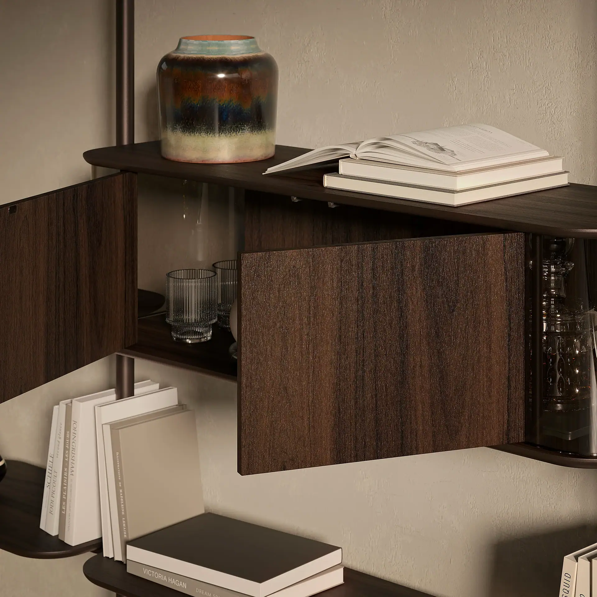 A dark wood wall-mounted shelving unit with open cabinet doors, displaying books, a multi-colored ceramic vase, glass tumblers, and a decanter against a light textured wall.
