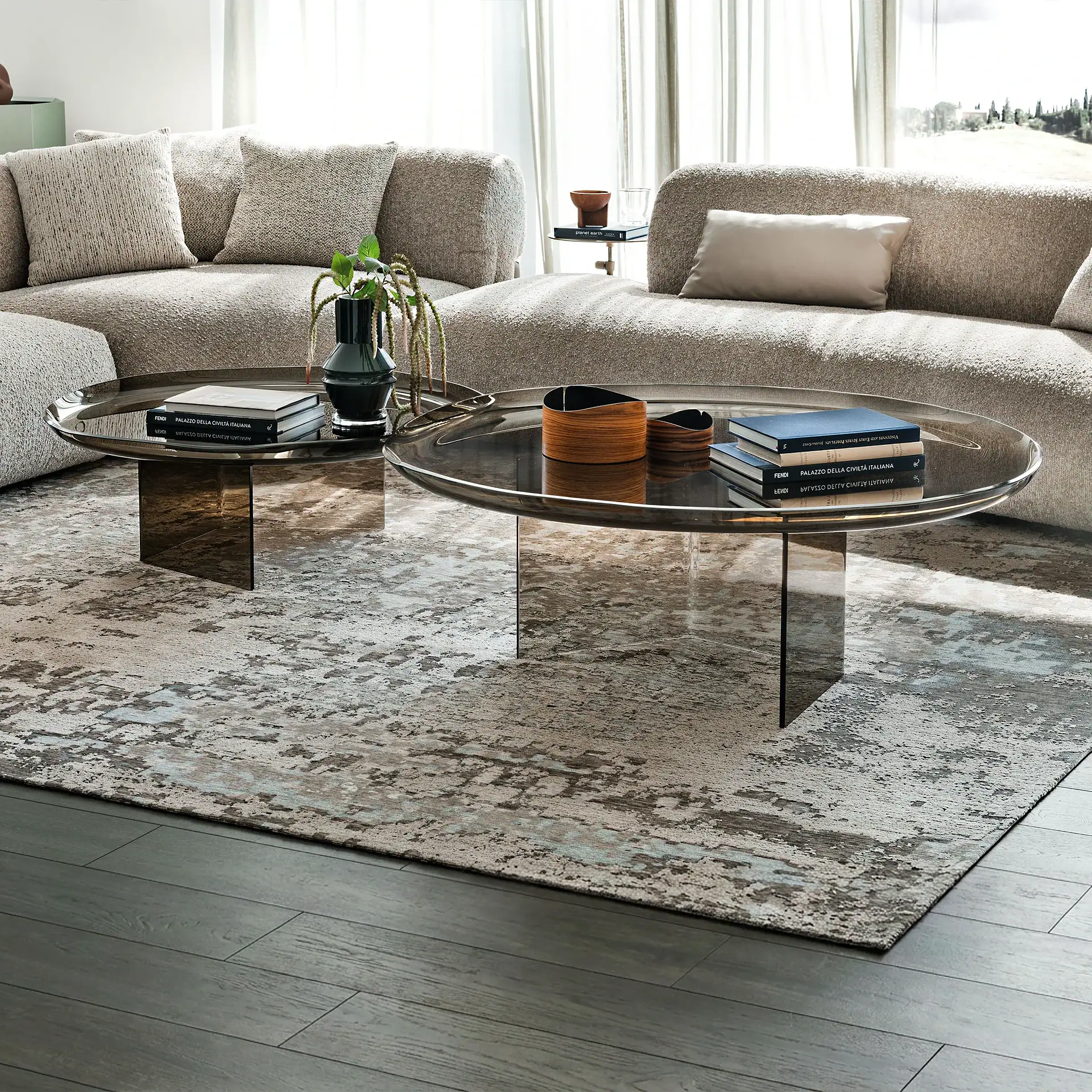 A modern living room with two curved, textured beige sofas, two round smoked glass coffee tables, and an abstract grey and blue rug on a dark wood floor. Natural light enters from large windows.