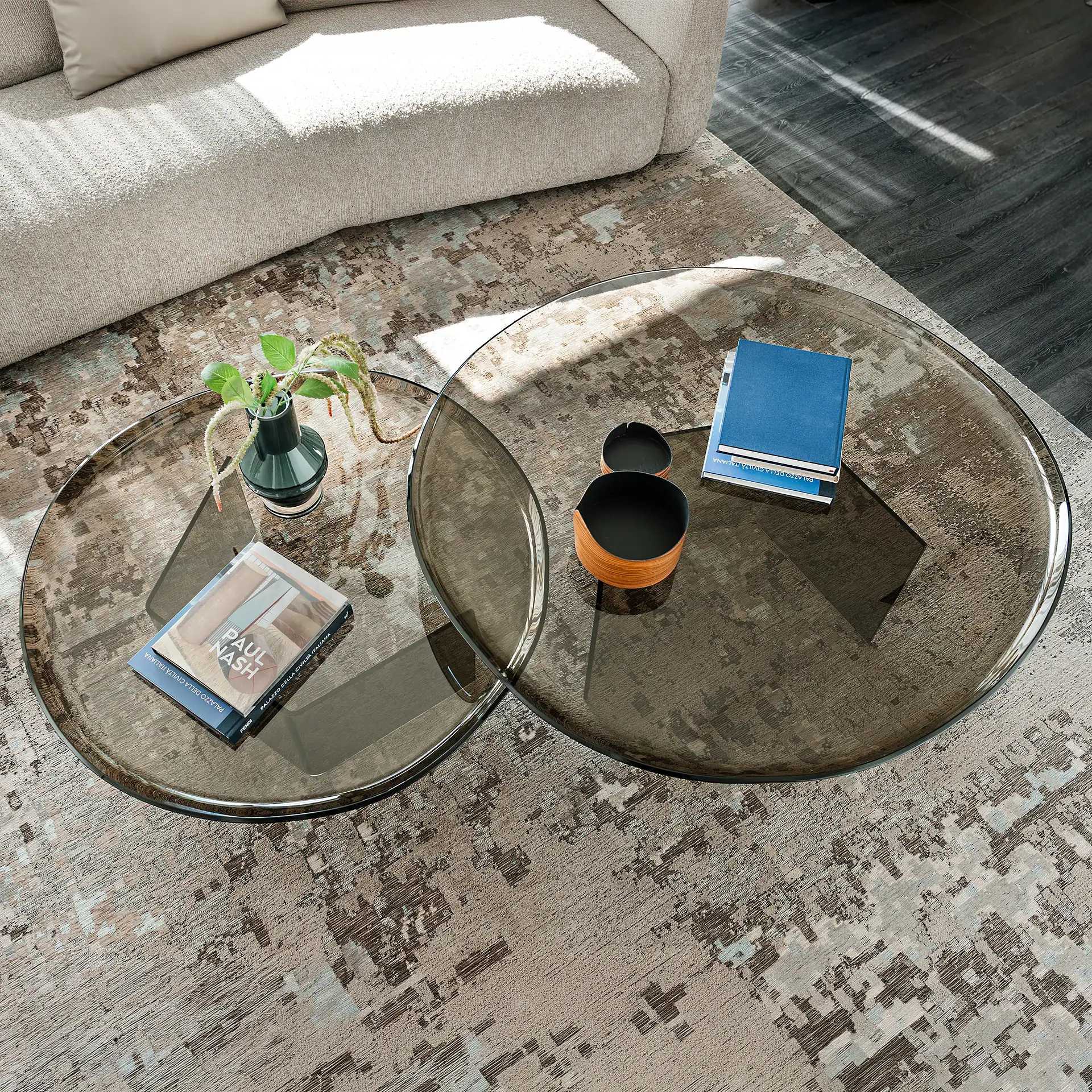 Overhead view of two overlapping round smoked glass coffee tables on an abstract patterned rug, with a beige sofa in the background. Tables hold books, a plant, and decorative bowls.