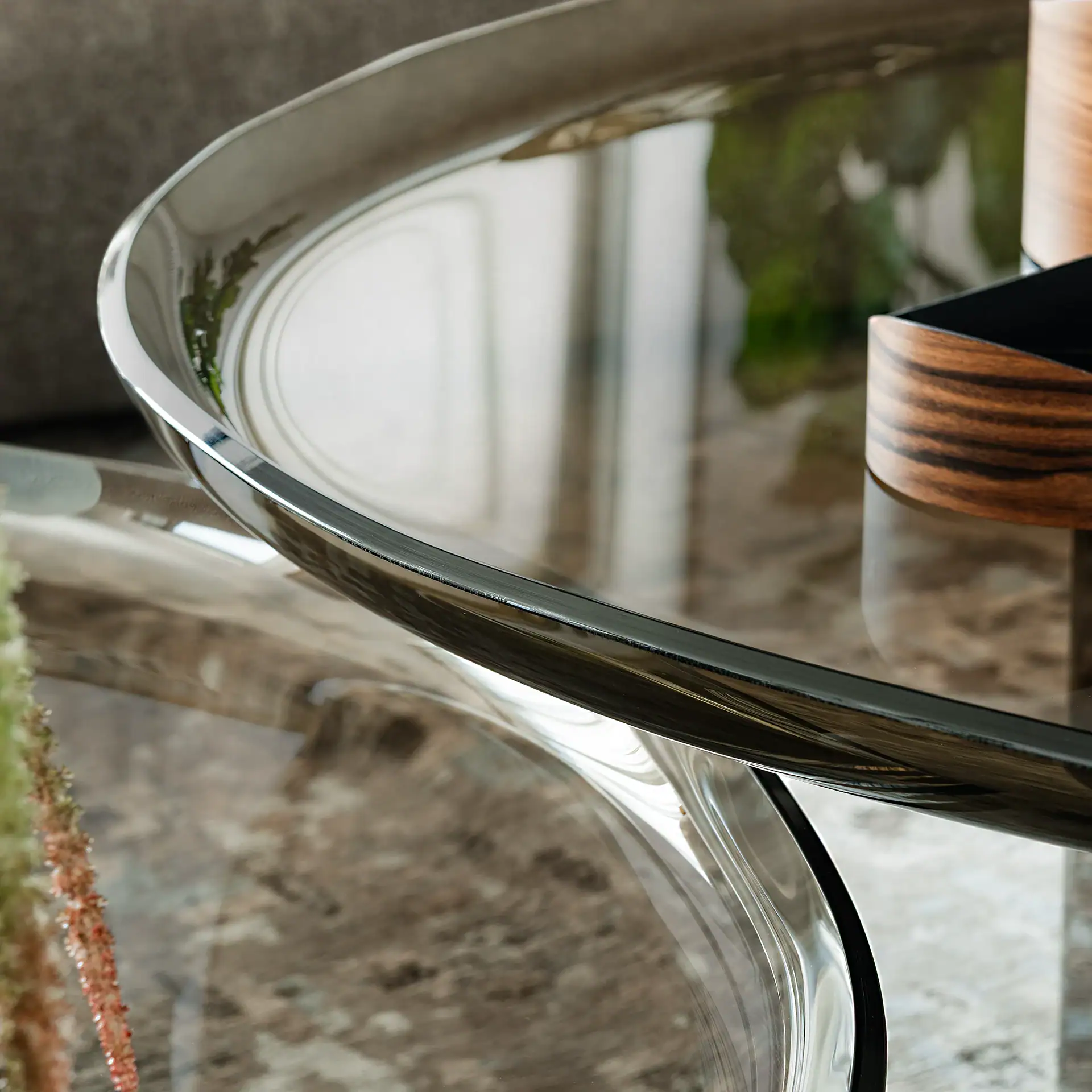 Close-up of a modern coffee table with a clear glass top, polished metallic rim, and clear curved base, reflecting a plant, floor, and a wooden cylindrical object.