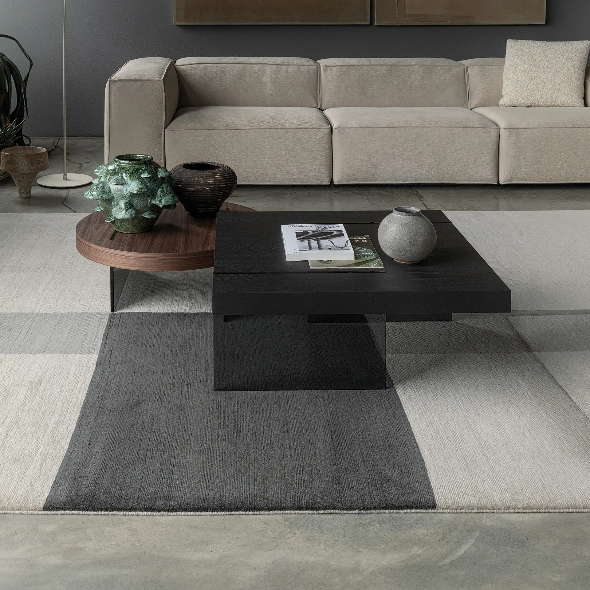 A modern living room with a beige modular sofa, dark coffee tables, and a geometric patterned rug on a concrete floor.